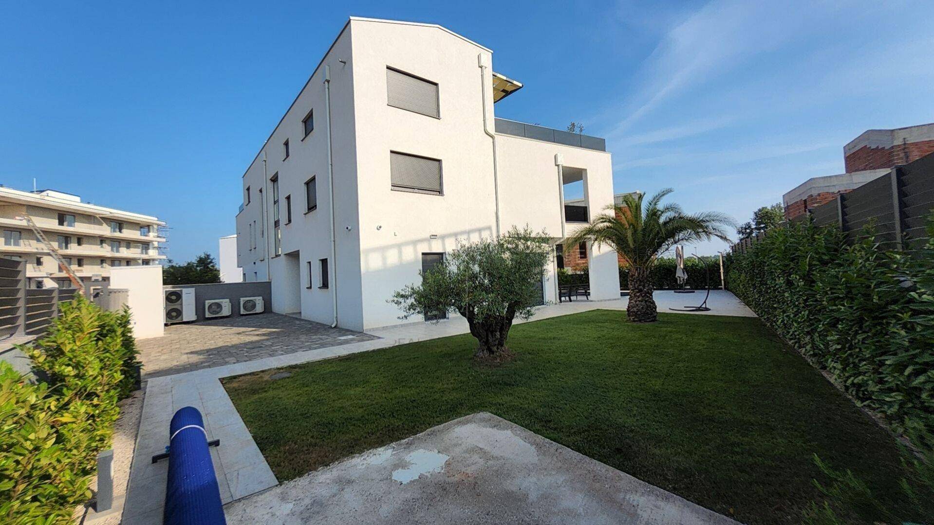 Moderne weiße Villa mit großem Garten, Palmen und gepflegter Rasenfläche unter blauem Himmel.