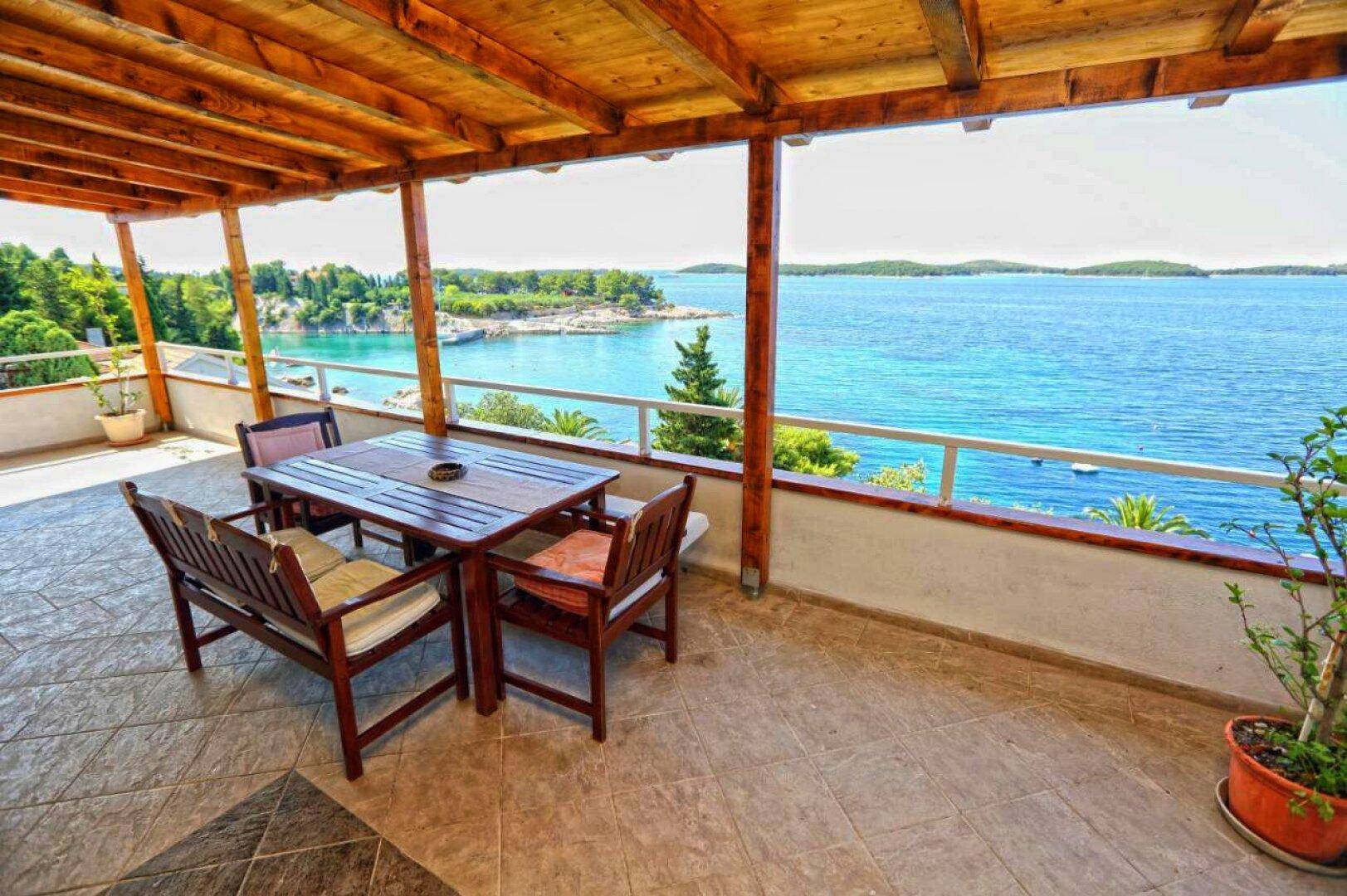 Überdachte Terrasse mit Holztisch und Stühlen, bietet einen weiten Blick auf das azurblaue Meer.