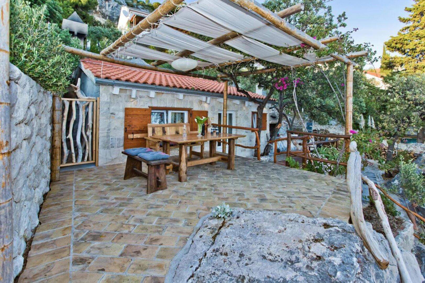 Gemütliche Terrasse mit Holztisch und Pergola, umgeben von mediterraner Vegetation.