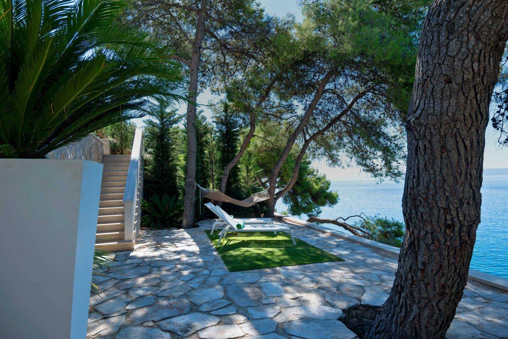 Idyllischer Gartenbereich mit Sonnenliege und Hängematte unter Bäumen, mit Blick auf das glitzernde Meer.
