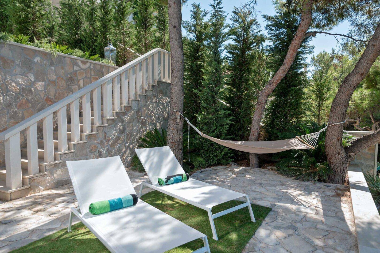Sonniger Gartenbereich mit bequemen Liegen und Hängematte, umgeben von hohen Bäumen und einer Steintreppe.