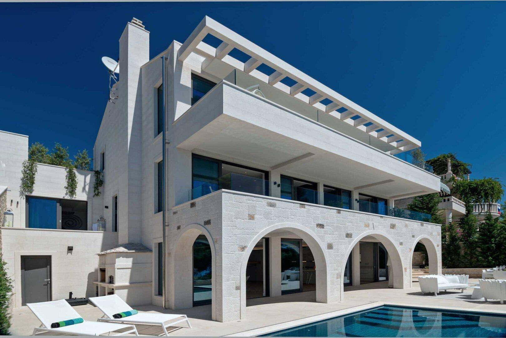 Moderne weiße Villa mit mehreren Balkonen und einem großen Poolbereich unter strahlend blauem Himmel.