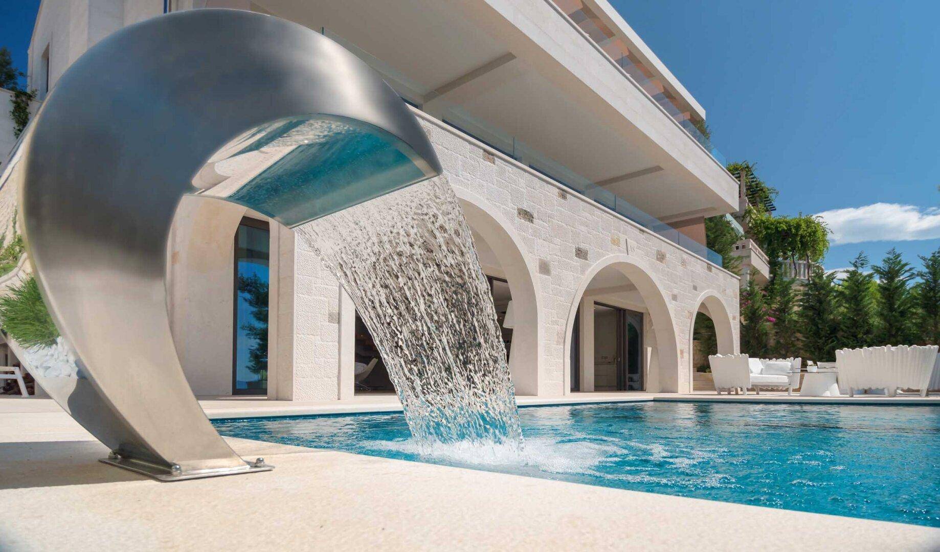 Moderner Poolbereich mit elegantem Wasserfall und luxuriöser Villa im Hintergrund unter blauem Himmel.