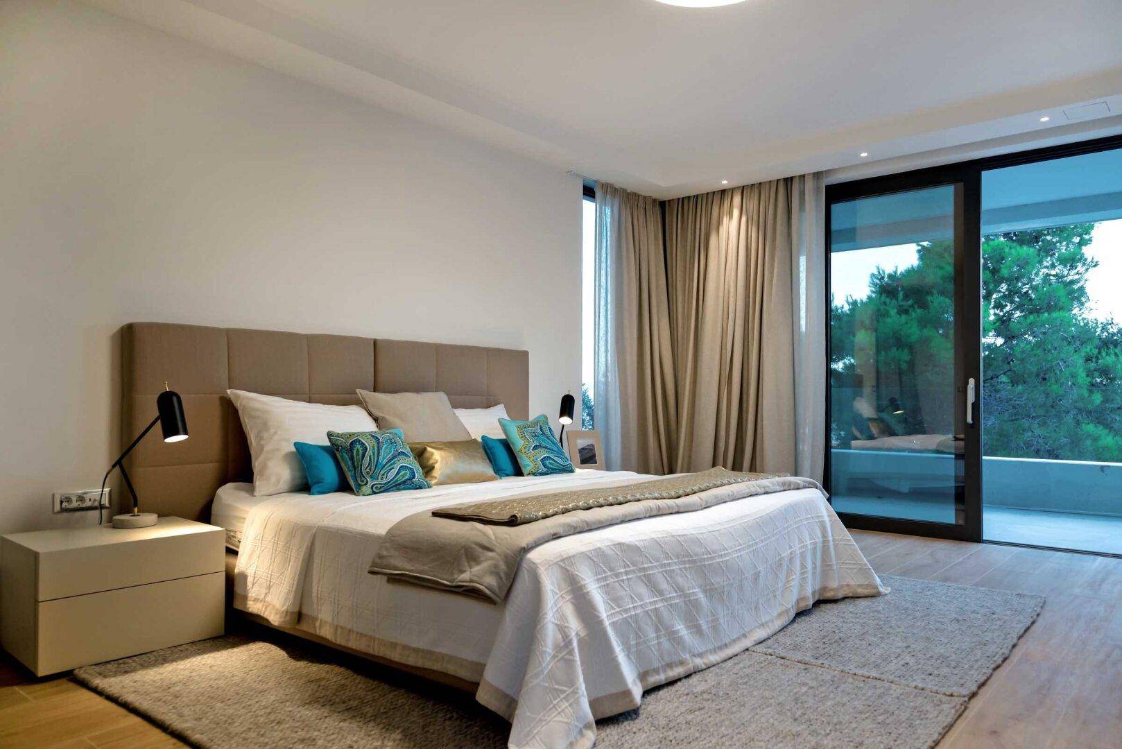 Modernes Schlafzimmer mit komfortablem Doppelbett, Holzboden und großen Fenstern mit Blick ins Grüne.