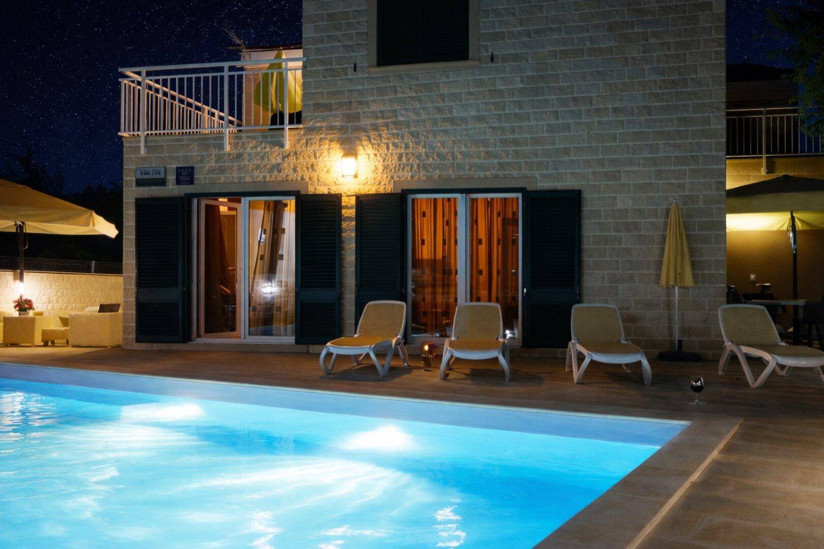 Nahaufnahme des beleuchteten Pools und der Terrasse mit Liegestühlen in der Nacht.