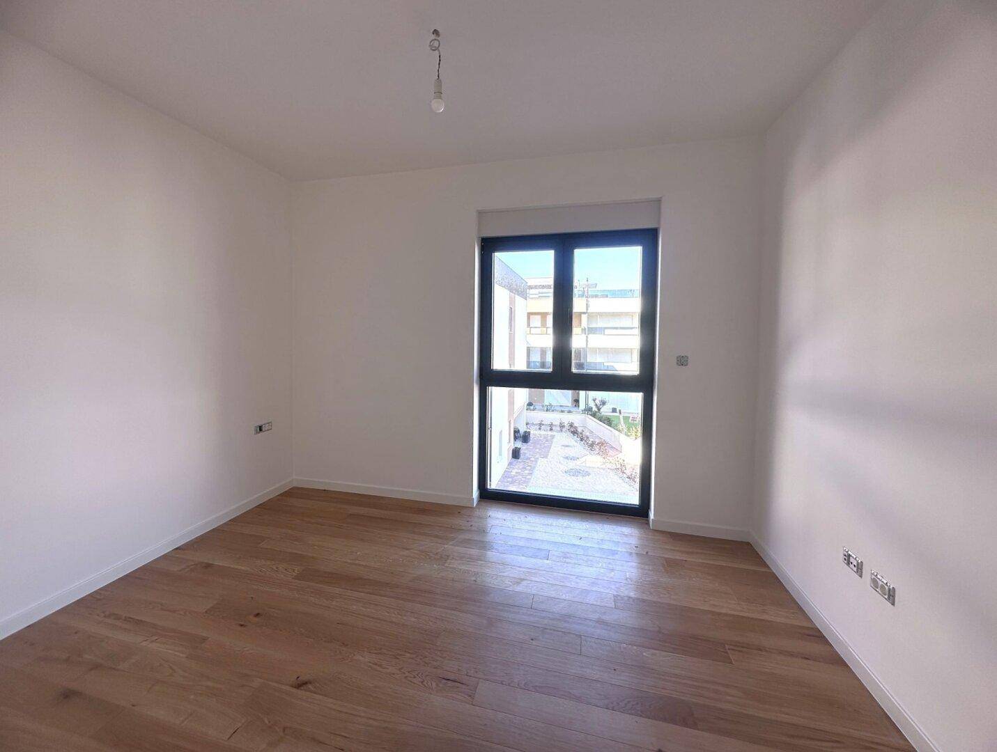 Schlafzimmer mit hellem Holzboden und großem Fenster mit Blick auf den Innenhof.
