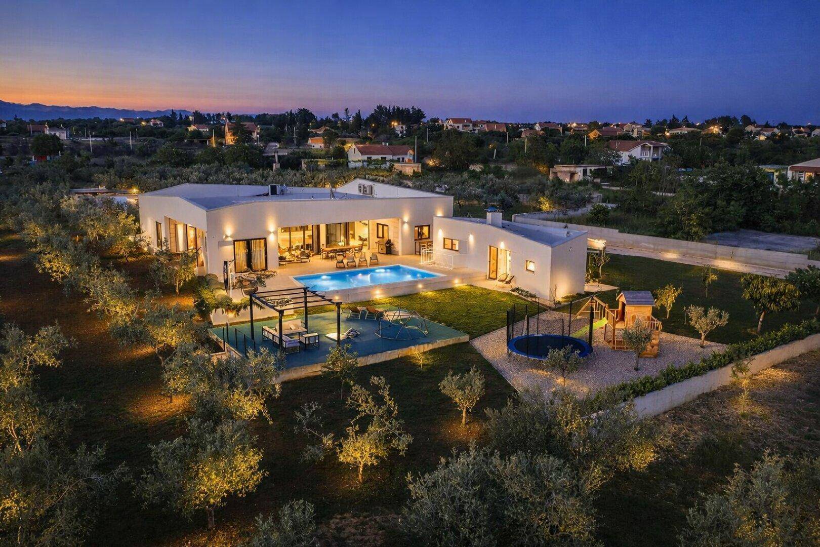 Luftaufnahme der modernen Villa mit Pool, Spielplatz und weitläufigem Garten bei Sonnenuntergang.