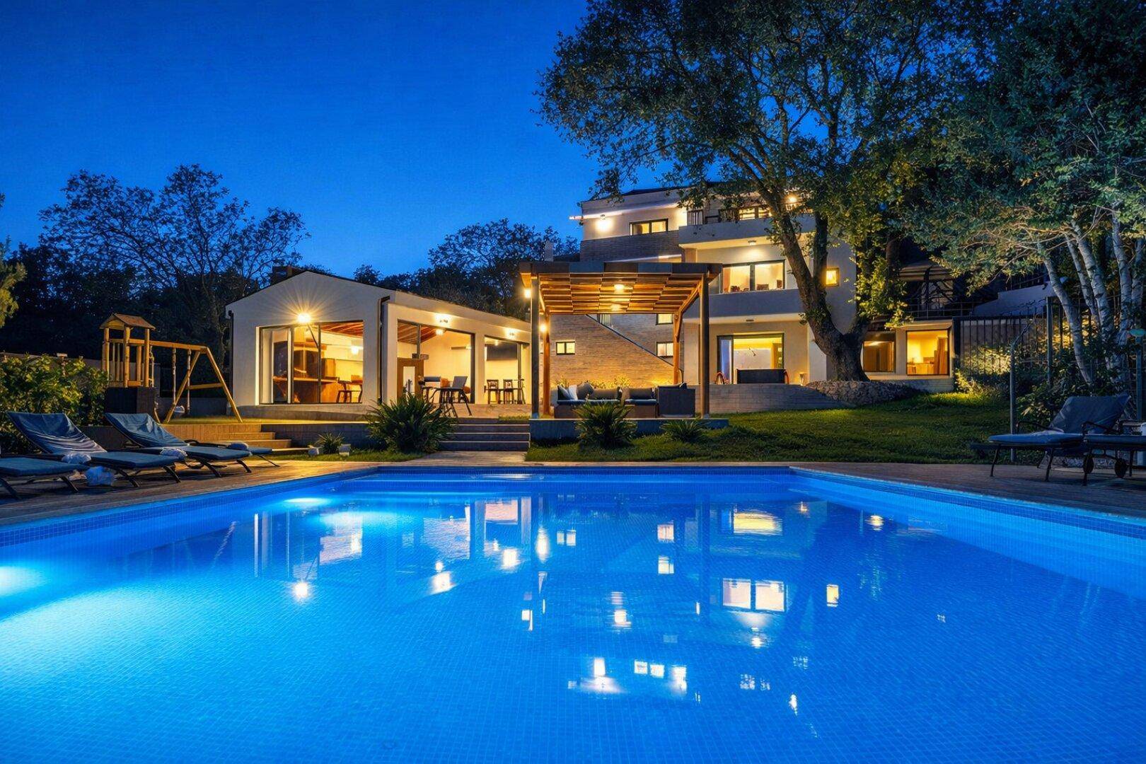 Großer beleuchteter Poolbereich mit Liegestühlen und Blick auf das moderne Haus bei Nacht.