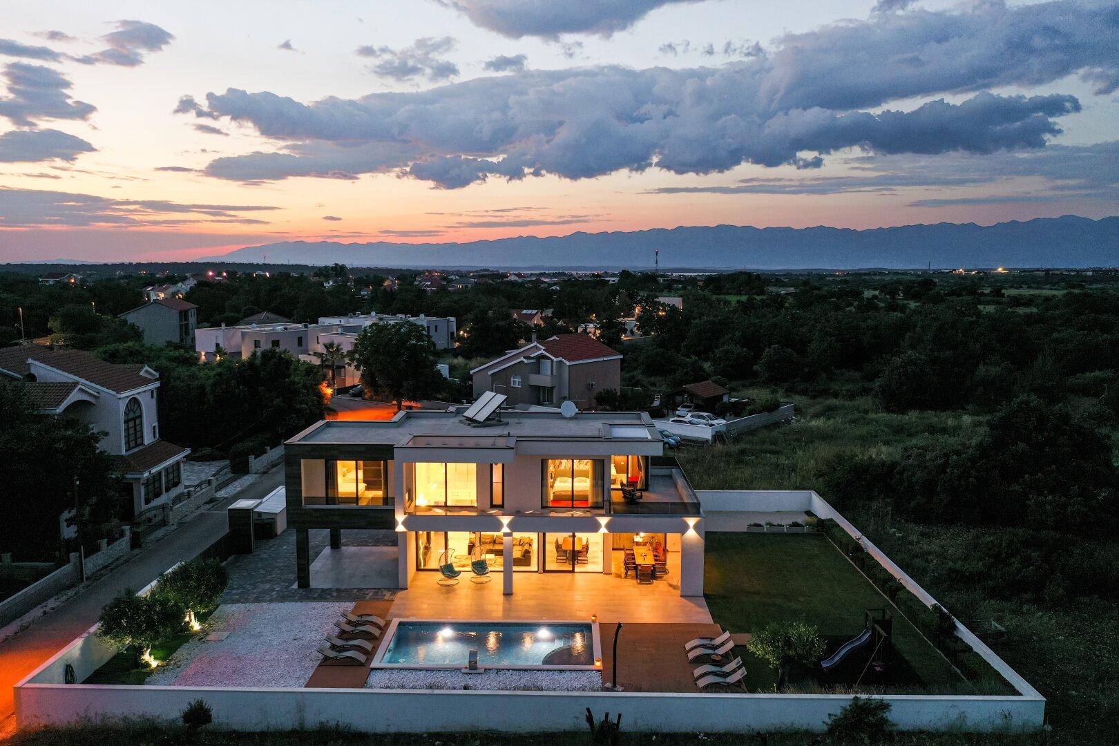 Luftaufnahme der modernen Villa mit beleuchtetem Poolbereich und umliegender Landschaft bei Sonnenuntergang.