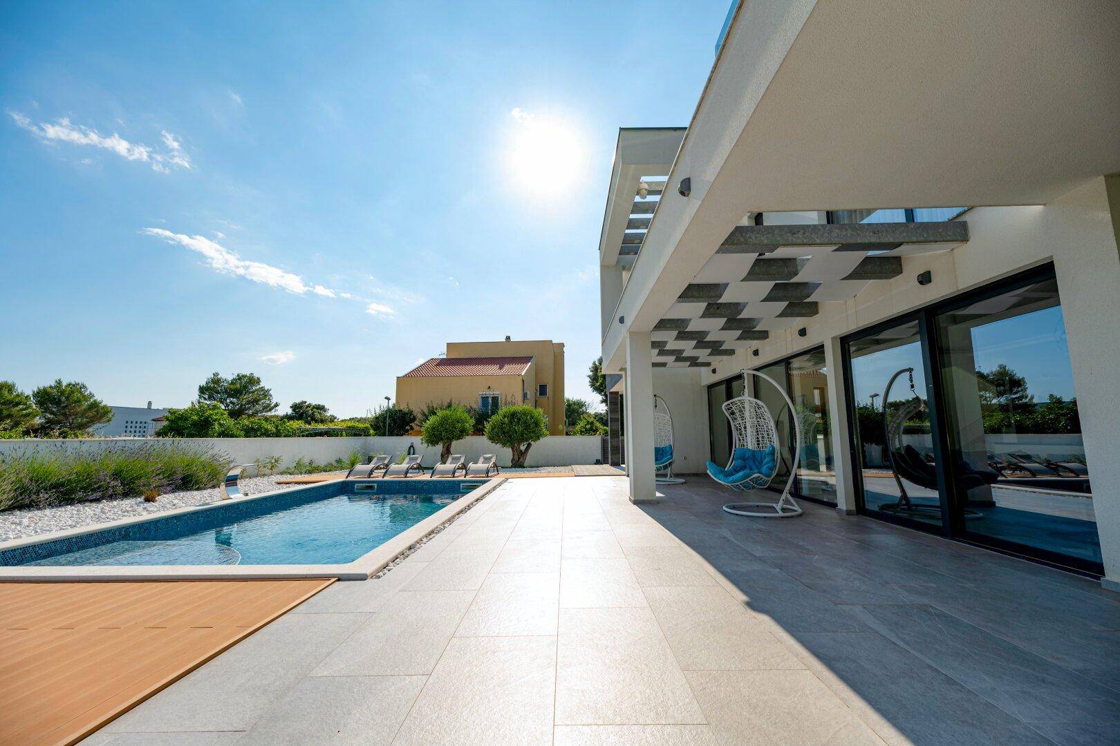 Sonniger Poolbereich mit Liegestühlen und moderner Architektur der Villa unter blauem Himmel.