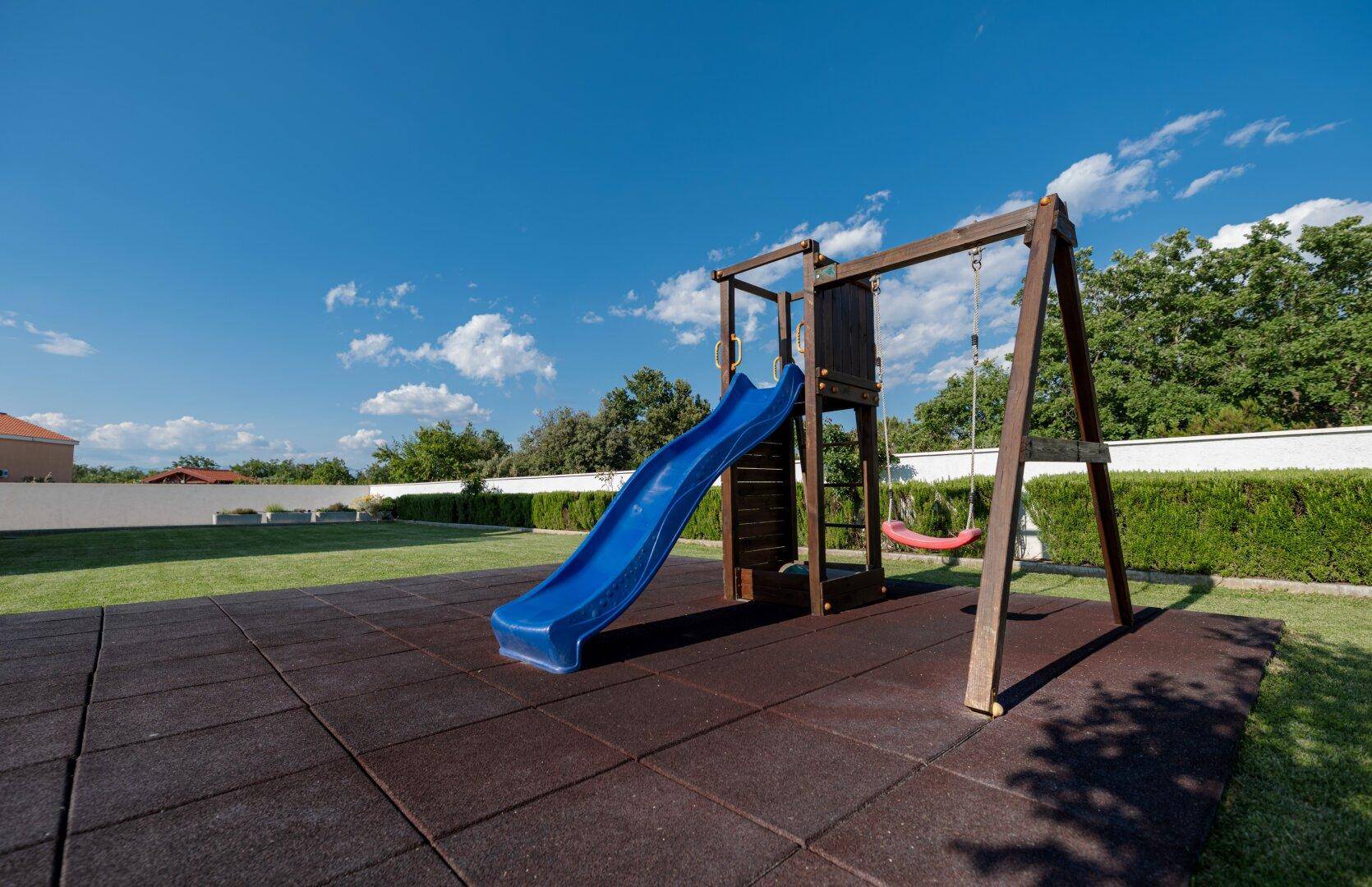 Grüner Gartenbereich mit Kinderspielplatz, Rutsche und Schaukel unter blauem Himmel.