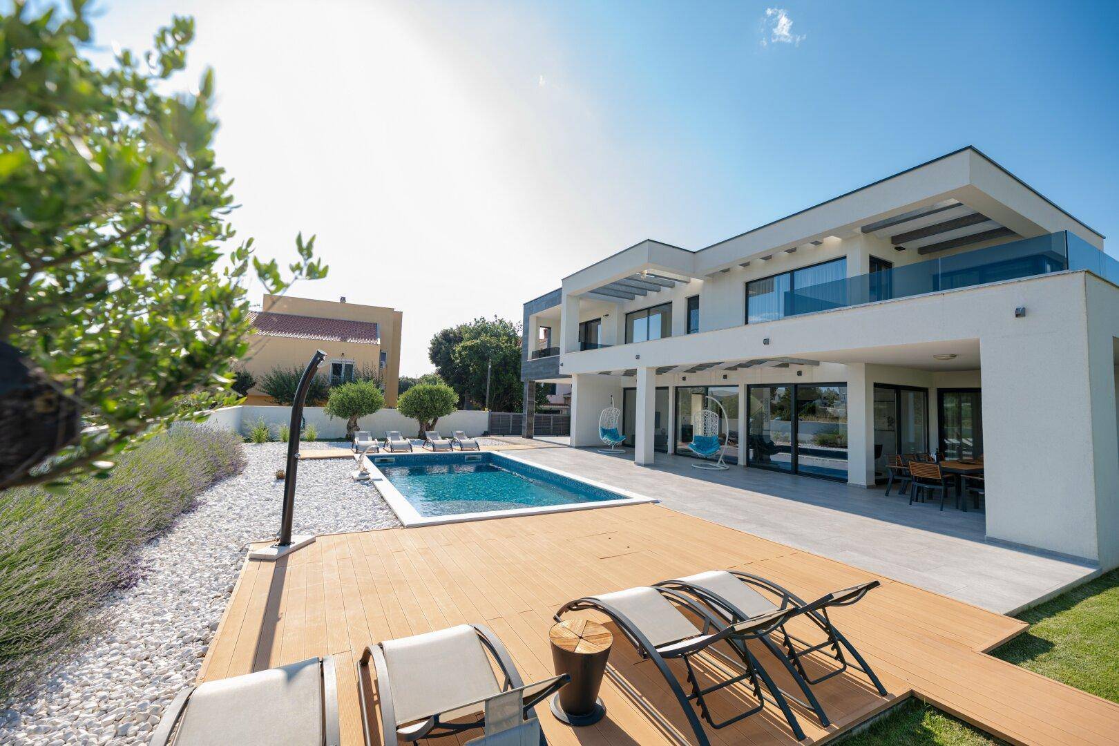 Weitläufiger Poolbereich mit Holzdeck, Liegestühlen und moderner Villa unter strahlendem Himmel.