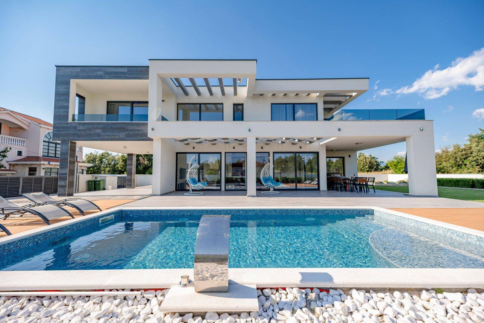 Moderne Villa am Tag mit großem Pool, Sonnenliegen und weitläufiger Terrasse unter blauem Himmel.