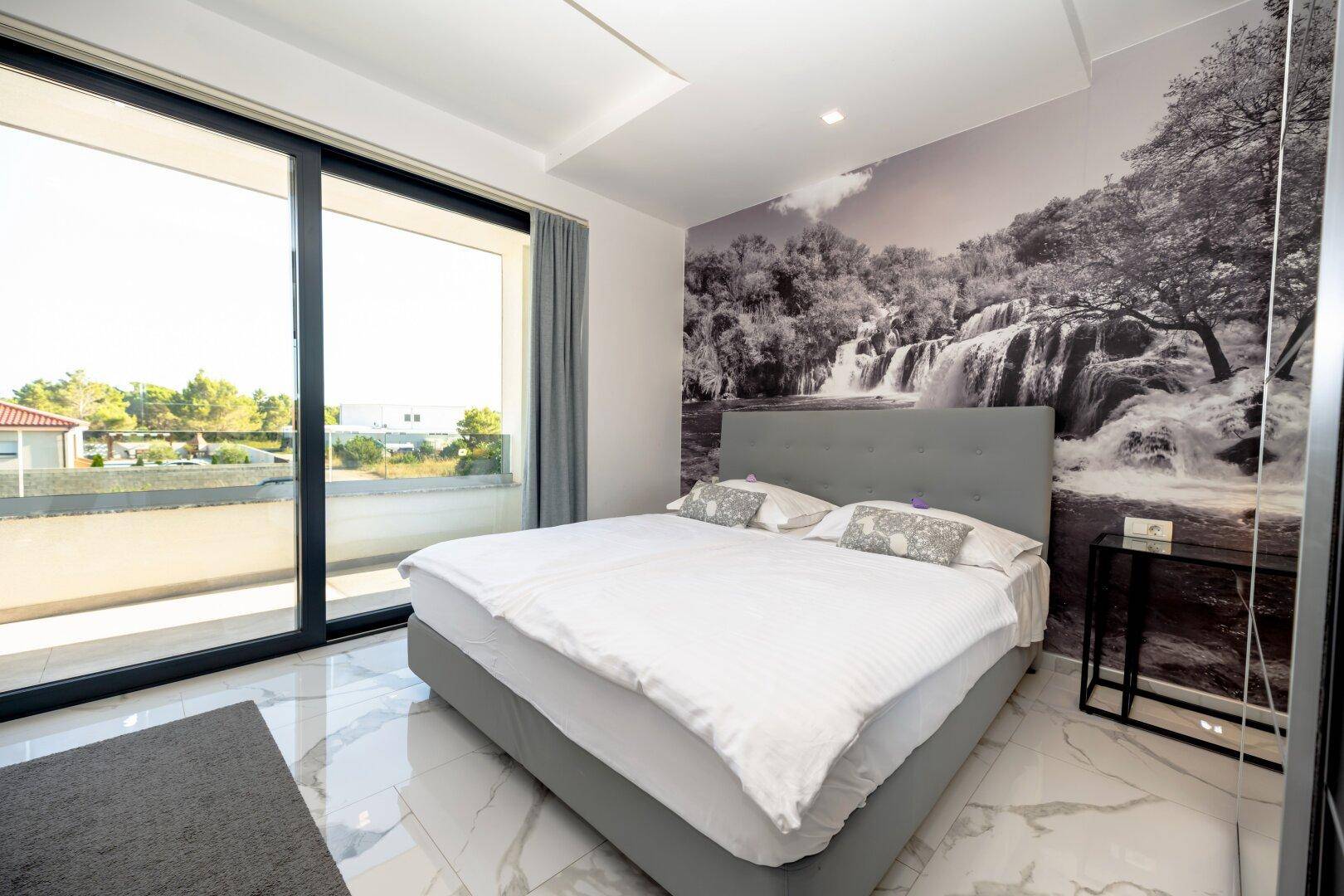 Modernes Schlafzimmer mit großem Bett, Wandbild und Zugang zu einem Balkon.