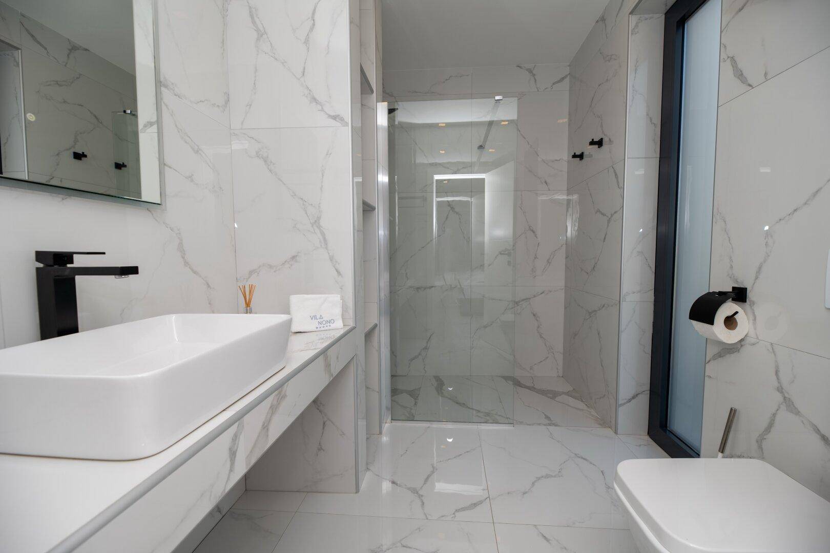 Luxuriöses Badezimmer mit weißen Marmorfliesen, ebenerdiger Dusche und modernem Waschtisch.
