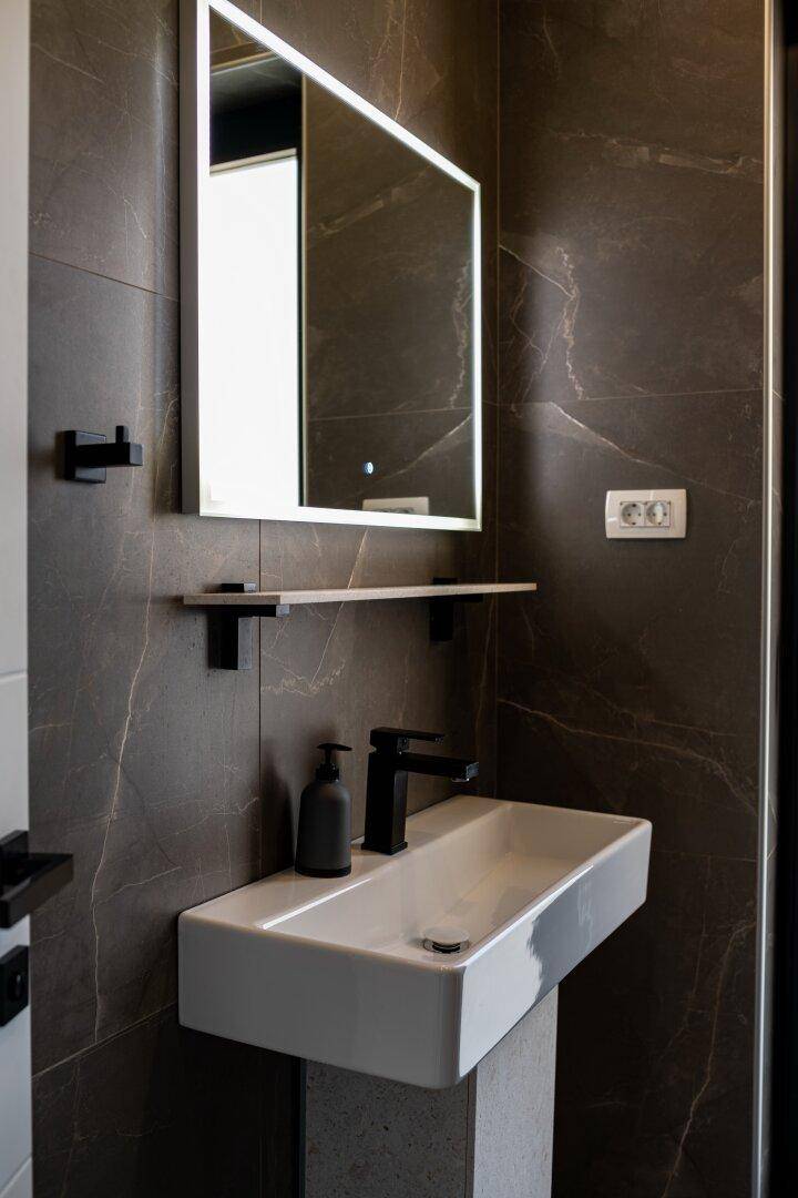 Modernes Badezimmer mit dunklen Fliesen, beleuchtetem Spiegel und stilvollem Waschtisch.