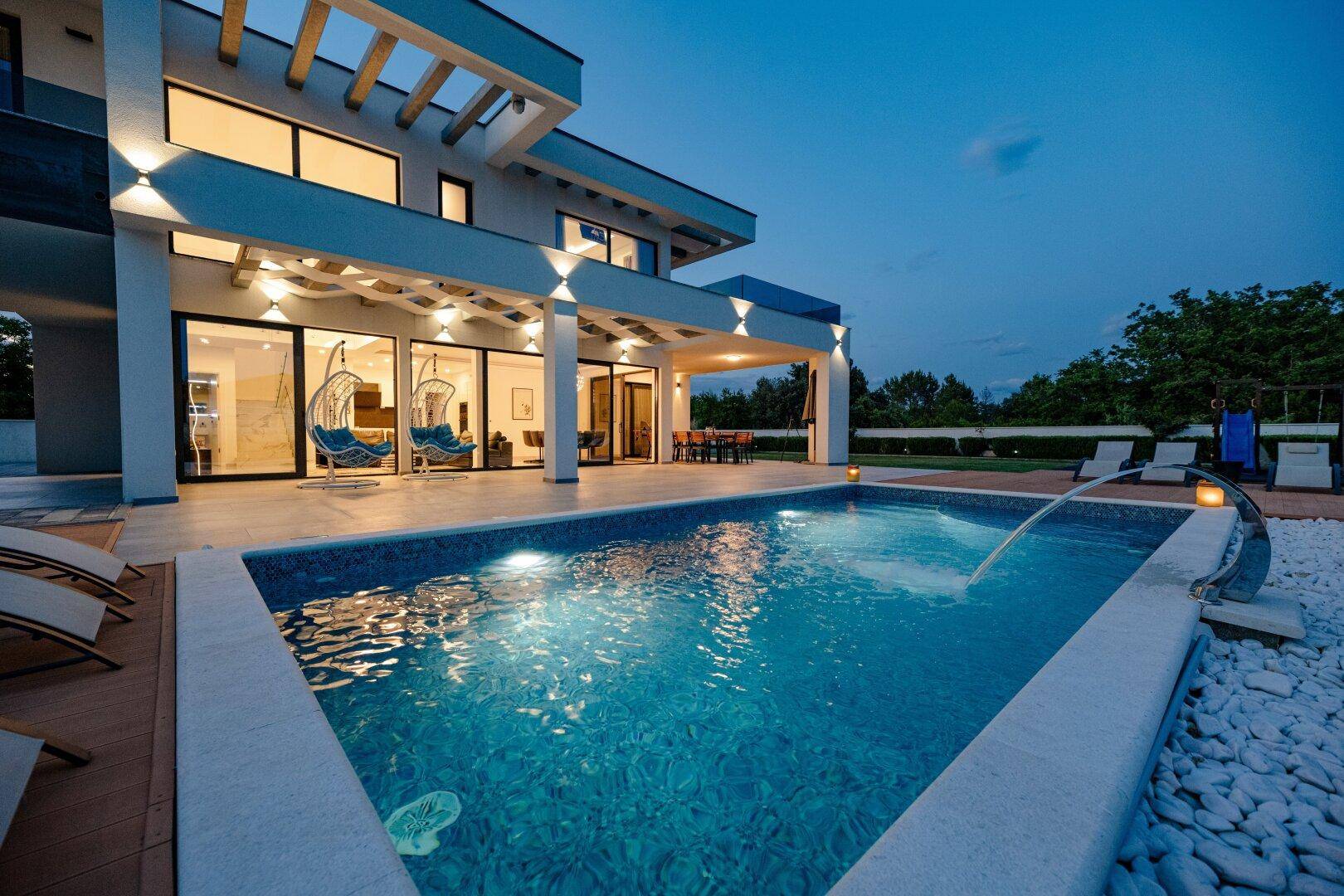 Beleuchteter Poolbereich der modernen Villa bei Nacht mit Wasserspiel und Liegestühlen.