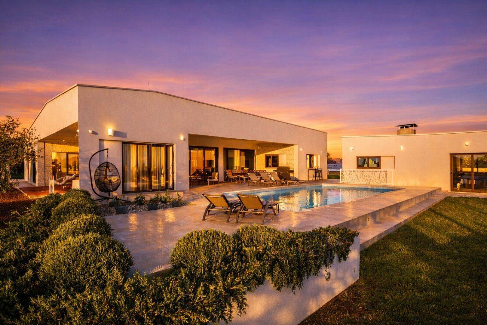 Moderne Villa mit großem Poolbereich, Sonnenliegen und einer Hängeschaukel bei Sonnenuntergang.