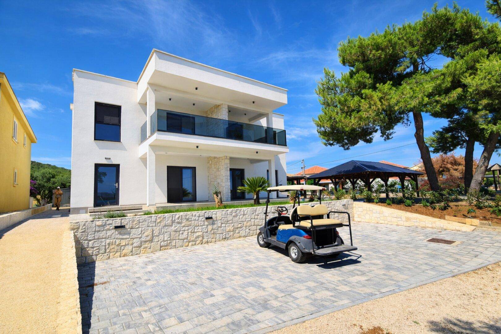 Moderne weiße Villa mit Parkplatz und Golfwagen vor dem Eingang unter blauem Himmel.