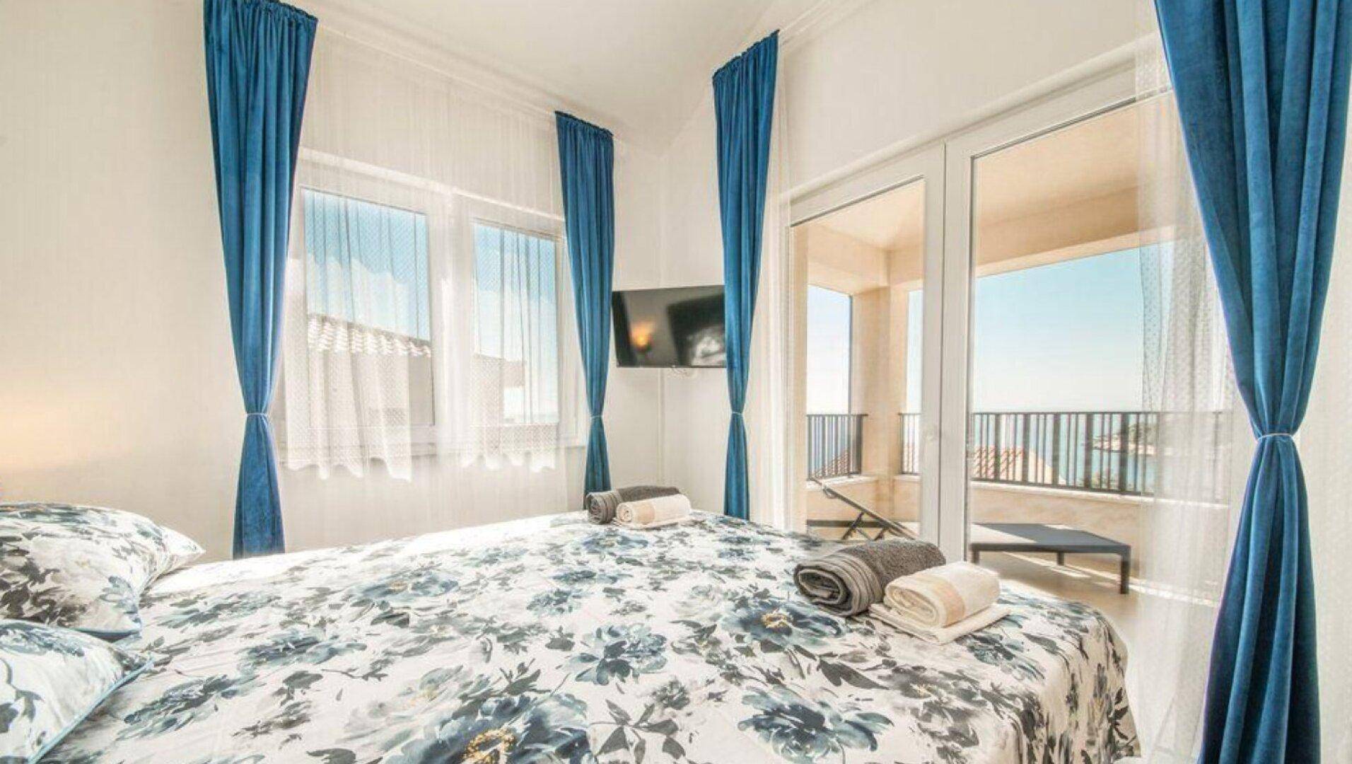 Helles Schlafzimmer mit Doppelbett, blauen Vorhängen und Zugang zum Balkon mit Meerblick.