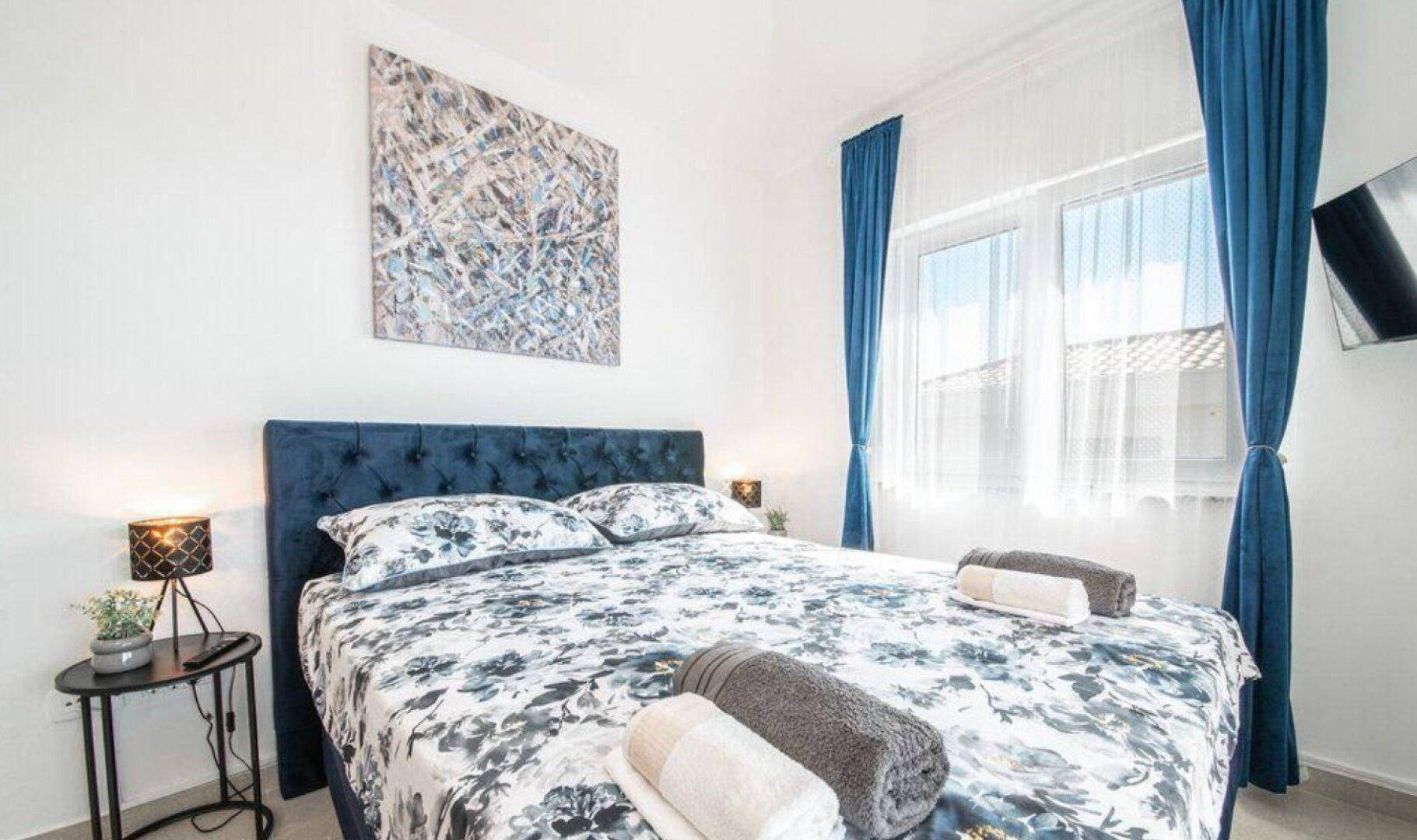 Schlafzimmer mit blauem Bett, floraler Bettwäsche und Fenster mit Blick nach draußen.