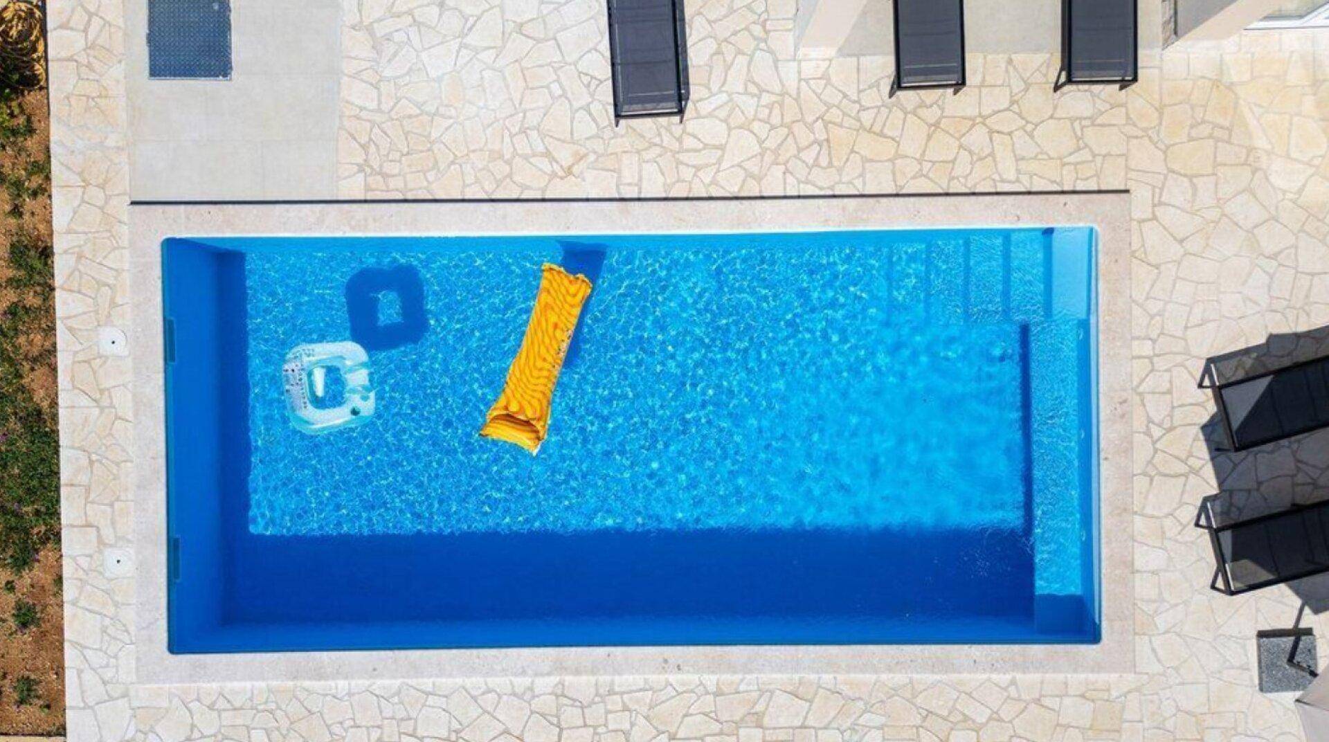 Vogelperspektive auf den rechteckigen Pool mit Liegen und Wasserspielzeug auf hellem Steinboden.