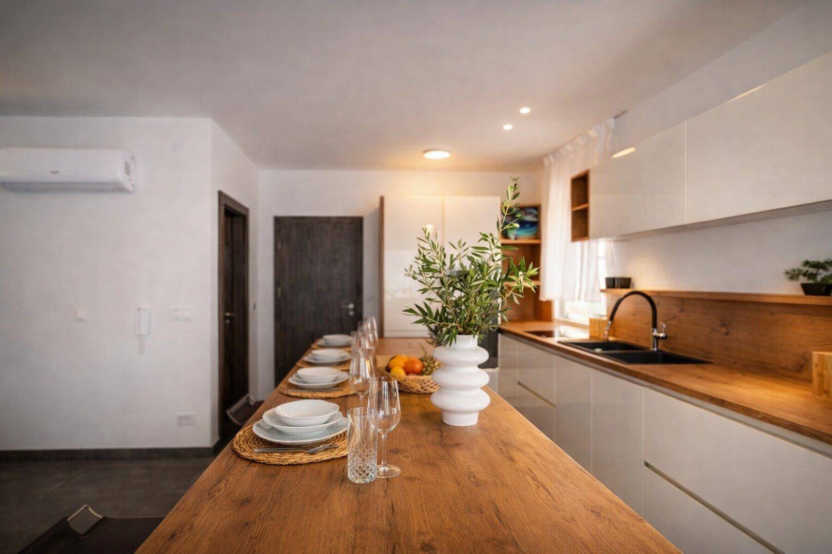 Helle Küche mit Holztheke, weißen Schränken und einem langen Esstisch mit Blick auf den Wohnbereich.