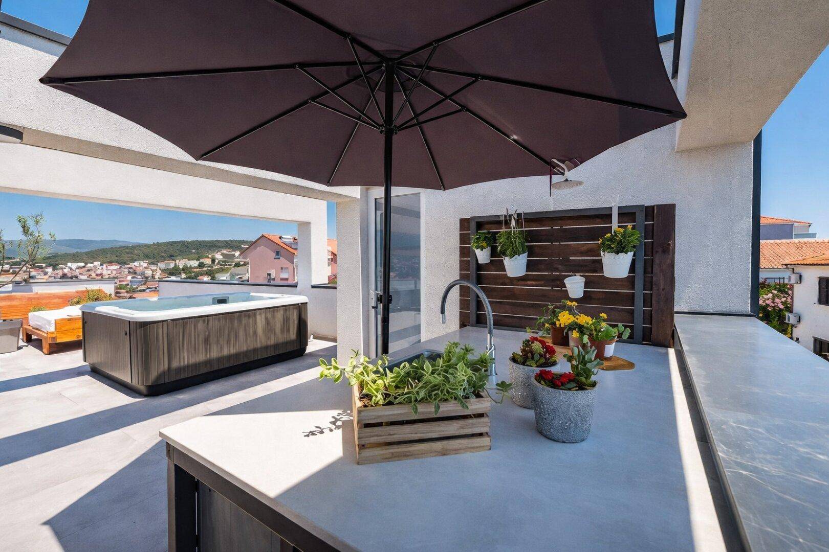 Dachterrasse mit Außenküche, Spüle und Whirlpool im Hintergrund unter einem Sonnenschirm.