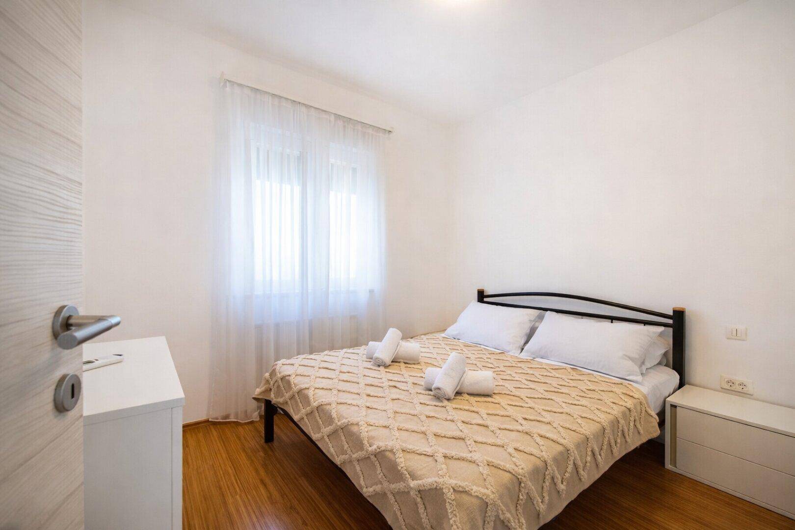 Helles Schlafzimmer mit Doppelbett, Holzboden und einem Fenster mit transparenten Vorhängen.