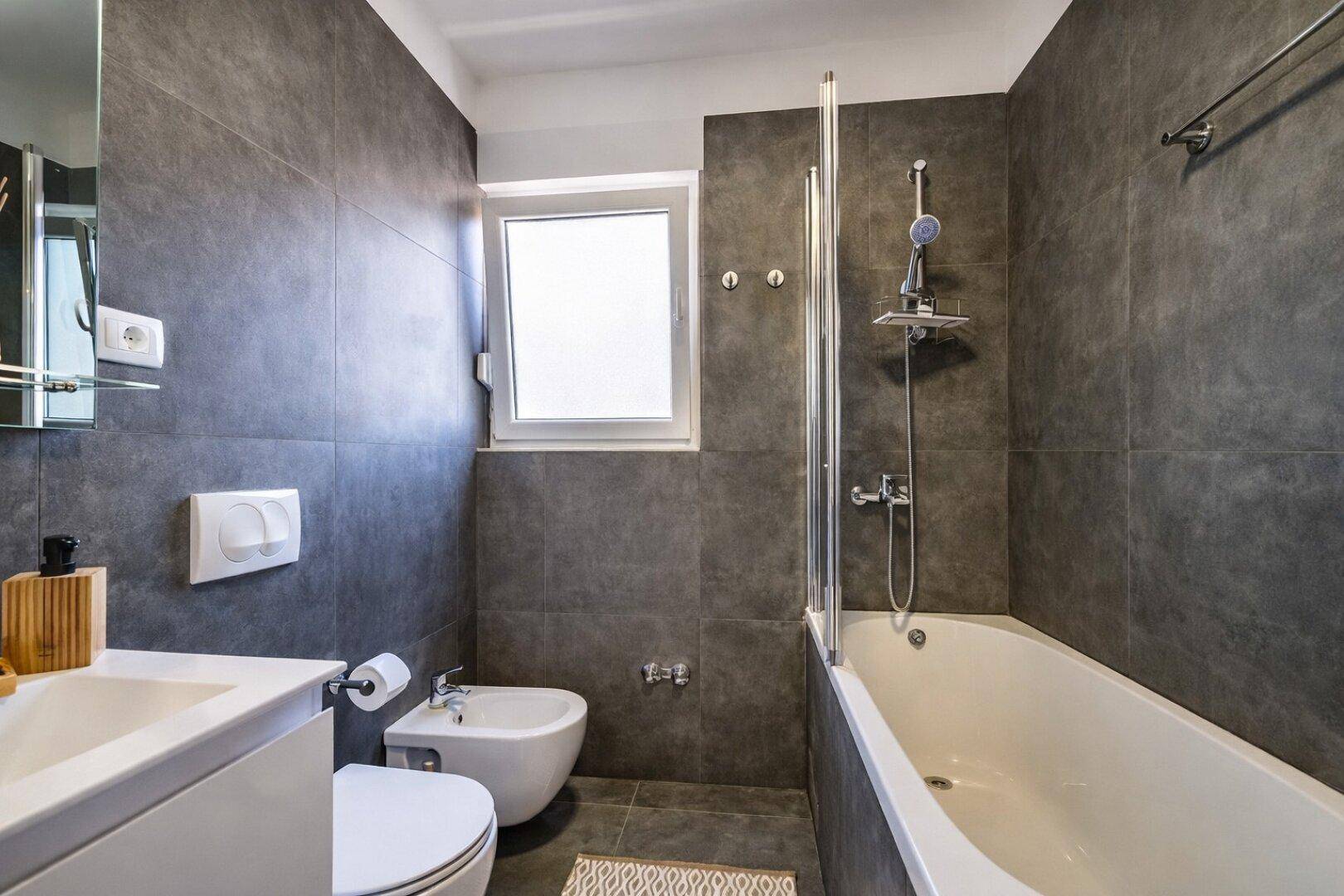 Badezimmer mit Badewanne, Dusche, Bidet und Fenster, ausgestattet mit dunklen Fliesen.