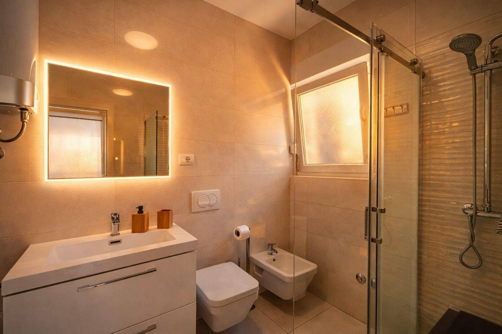 Modernes Badezimmer mit beleuchtetem Spiegel, Dusche, Bidet und hellen Fliesen.