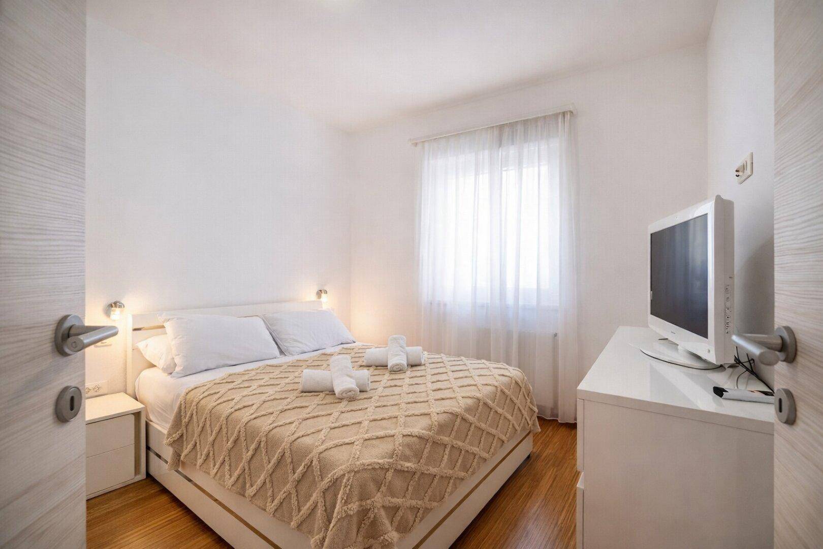 Gemütliches Schlafzimmer mit Doppelbett, Holzboden und einem Fenster mit hellen Vorhängen.