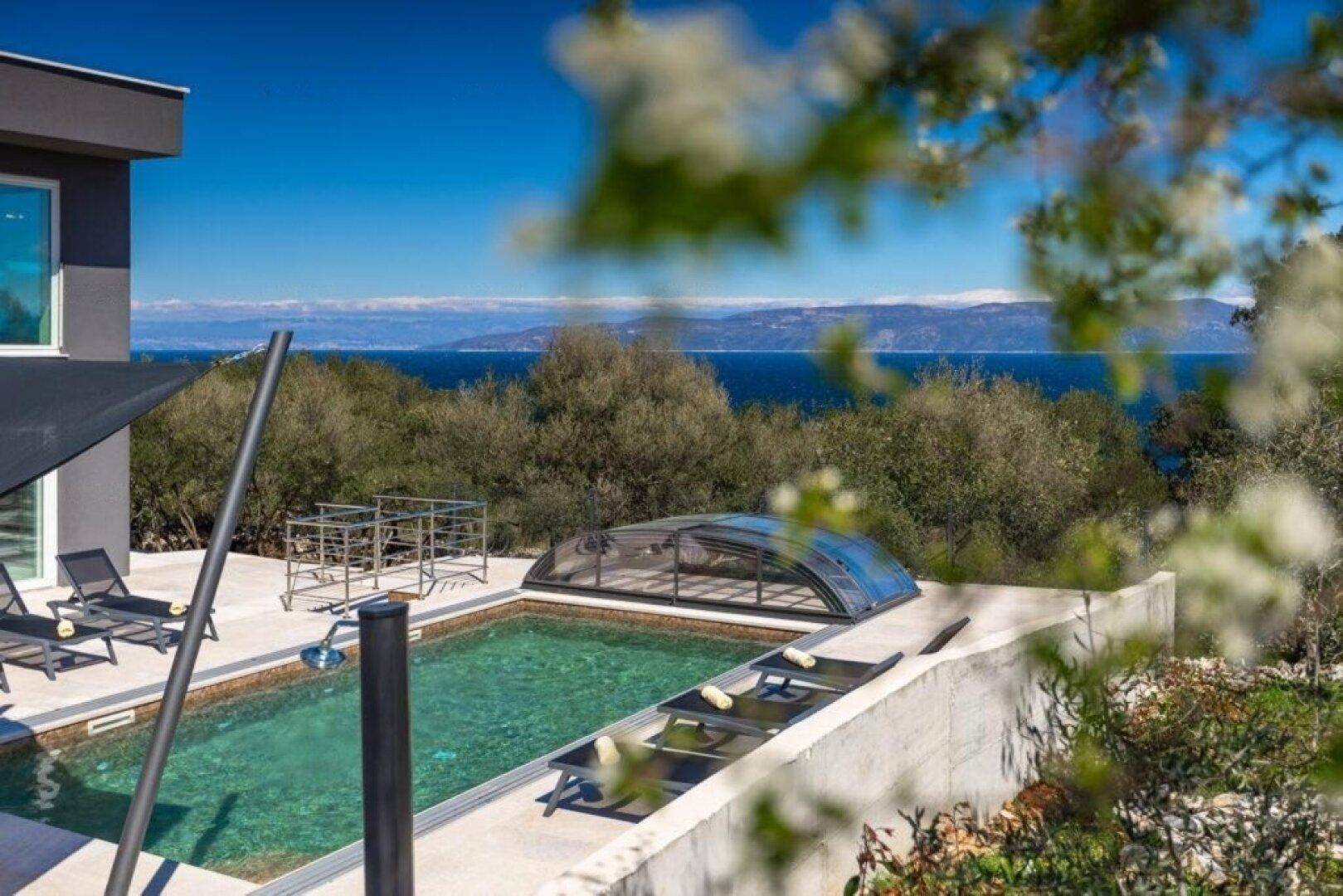 Poolbereich der Villa mit Meerblick und Bergen im Hintergrund unter strahlend blauem Himmel.