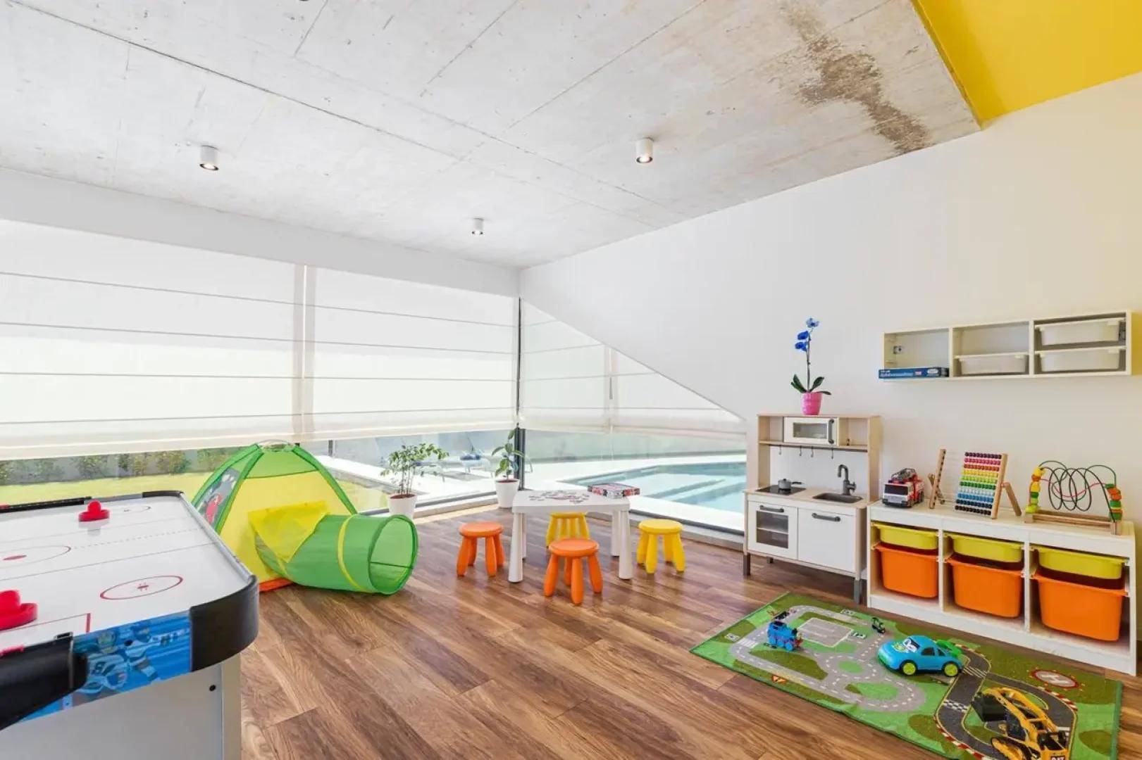 Helles Spielzimmer mit verschiedenen Spielsachen, einem Tisch und großen Fenstern mit Poolblick.