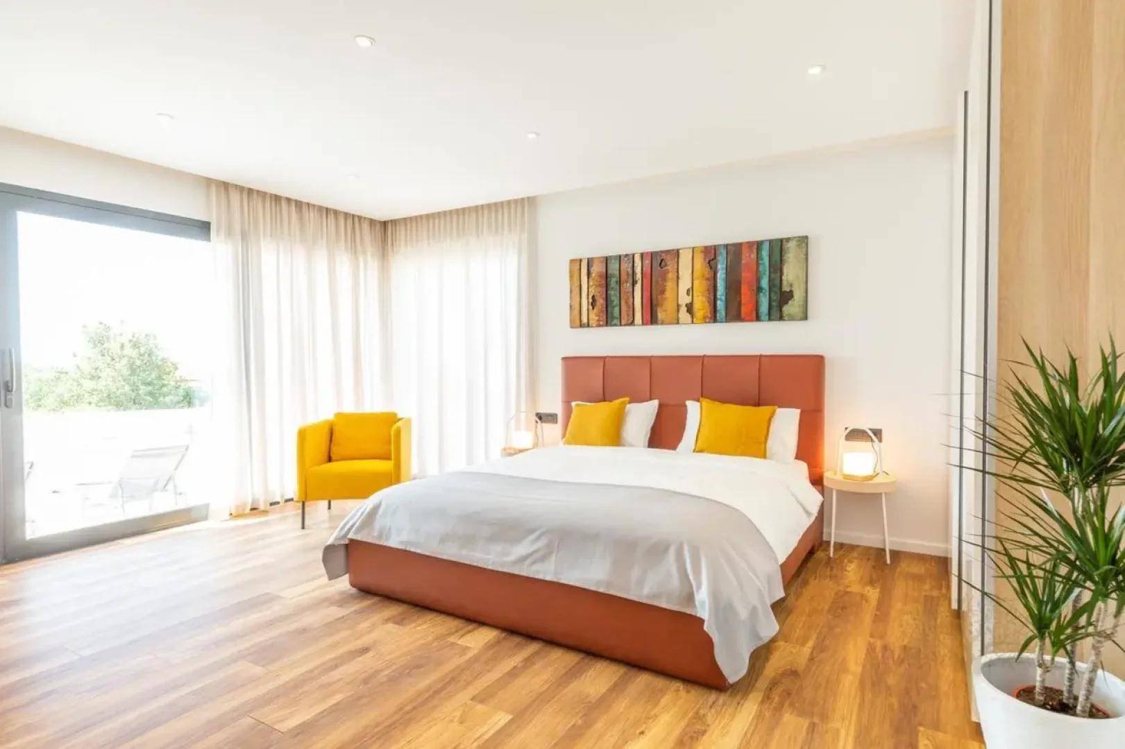 Helles Schlafzimmer mit großem Bett, gelbem Sessel und direktem Zugang zu einem Balkon oder einer Terrasse.