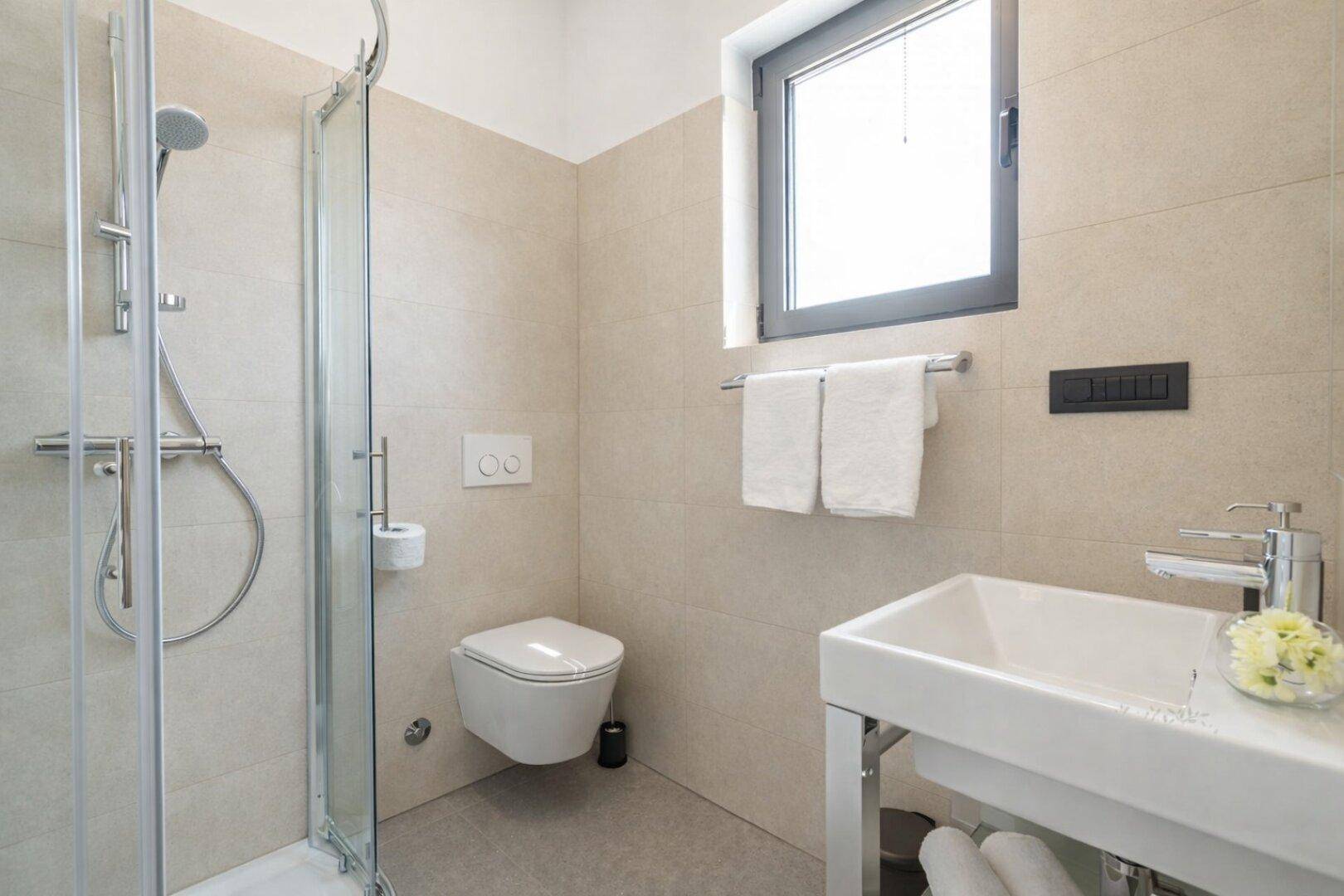 Helles Badezimmer mit Fenster, Duschkabine, wandhängendem WC und Einzelwaschbecken.