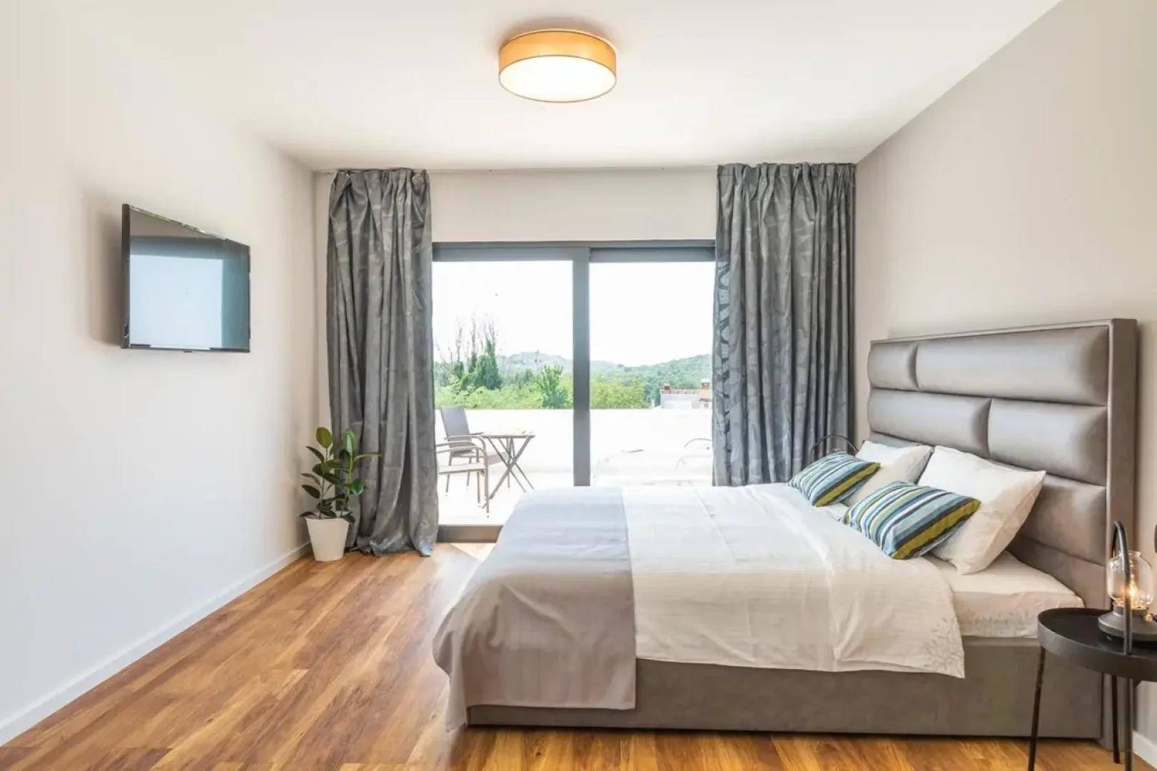 Komfortables Schlafzimmer mit Doppelbett, Wandfernseher und Zugang zu einem sonnigen Balkon.