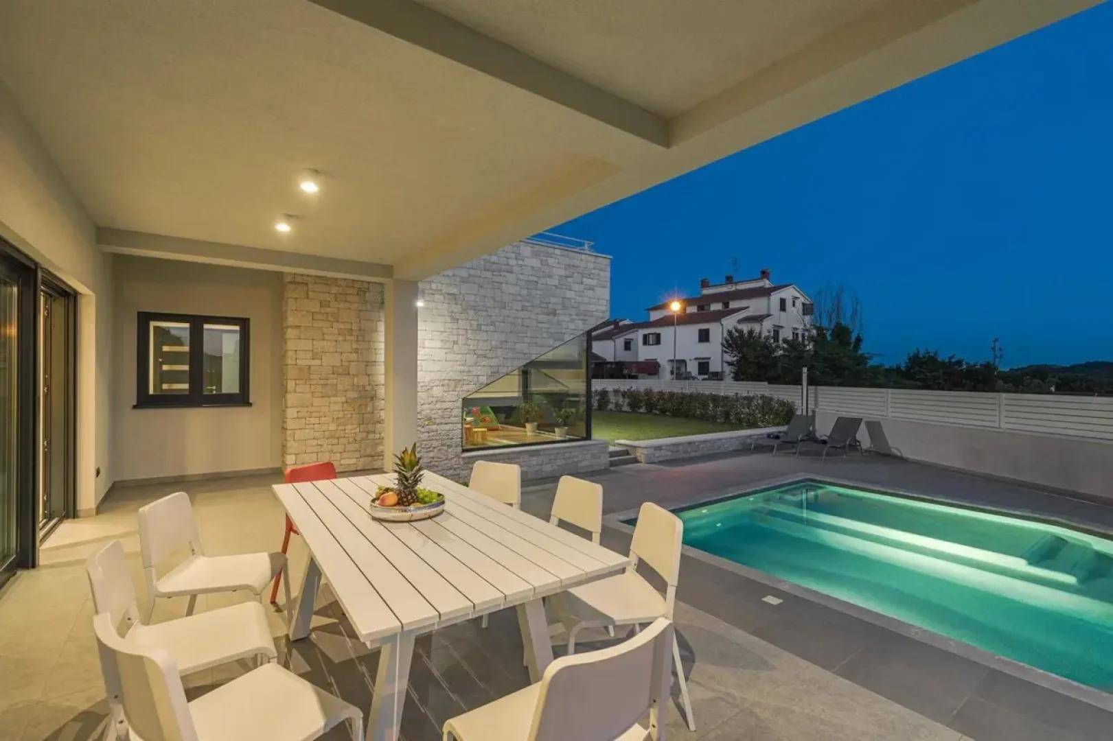 Überdachte Terrasse mit Esstisch und Stühlen direkt neben dem beleuchteten Swimmingpool am Abend.