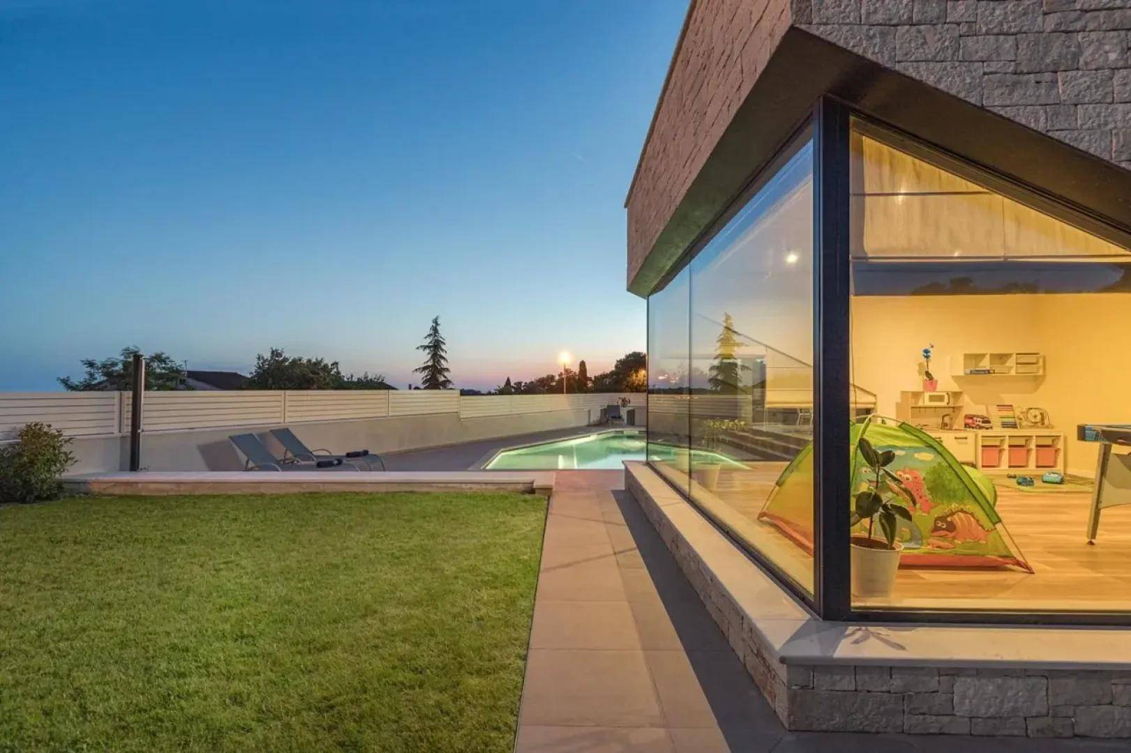 Seitliche Ansicht des Hauses mit Poolbereich und großem Glasfenster, das den Innenbereich beleuchtet.