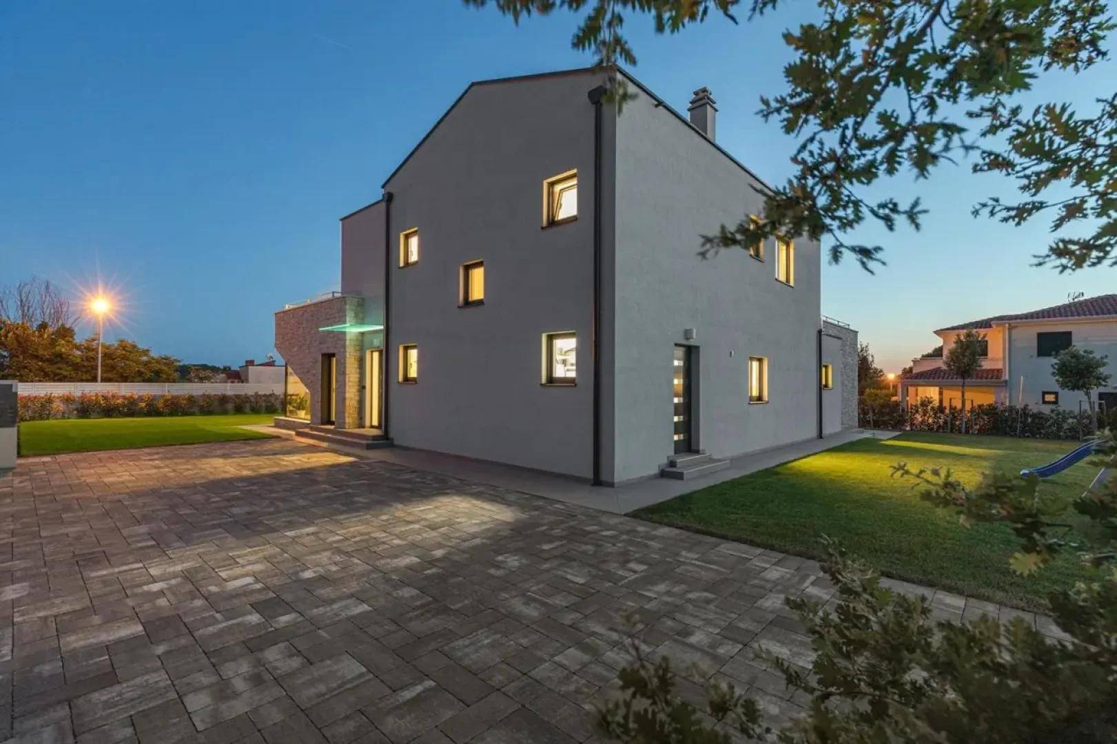 Seitenansicht des Hauses in der Abenddämmerung mit gepflasterter Auffahrt und beleuchtetem Gartenbereich.