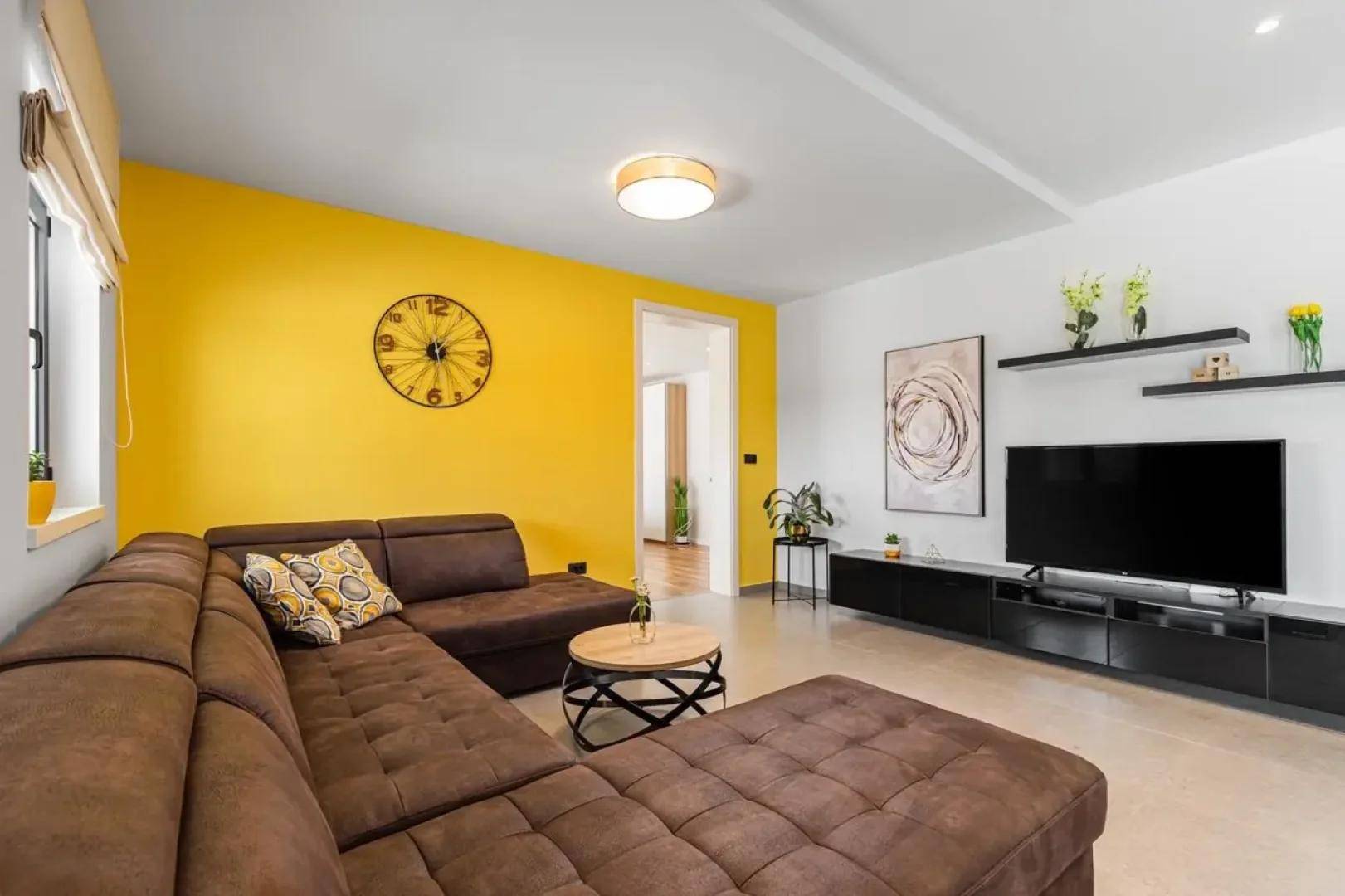 Gemütliches Wohnzimmer mit braunem Ecksofa, TV-Möbeln und einer lebhaften gelben Akzentwand.