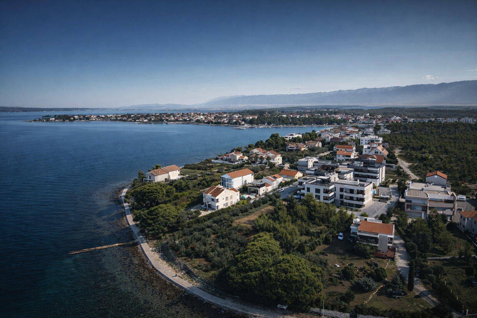 Luftaufnahme einer Küstensiedlung mit Häusern, viel Grün und dem Meer im Hintergrund.