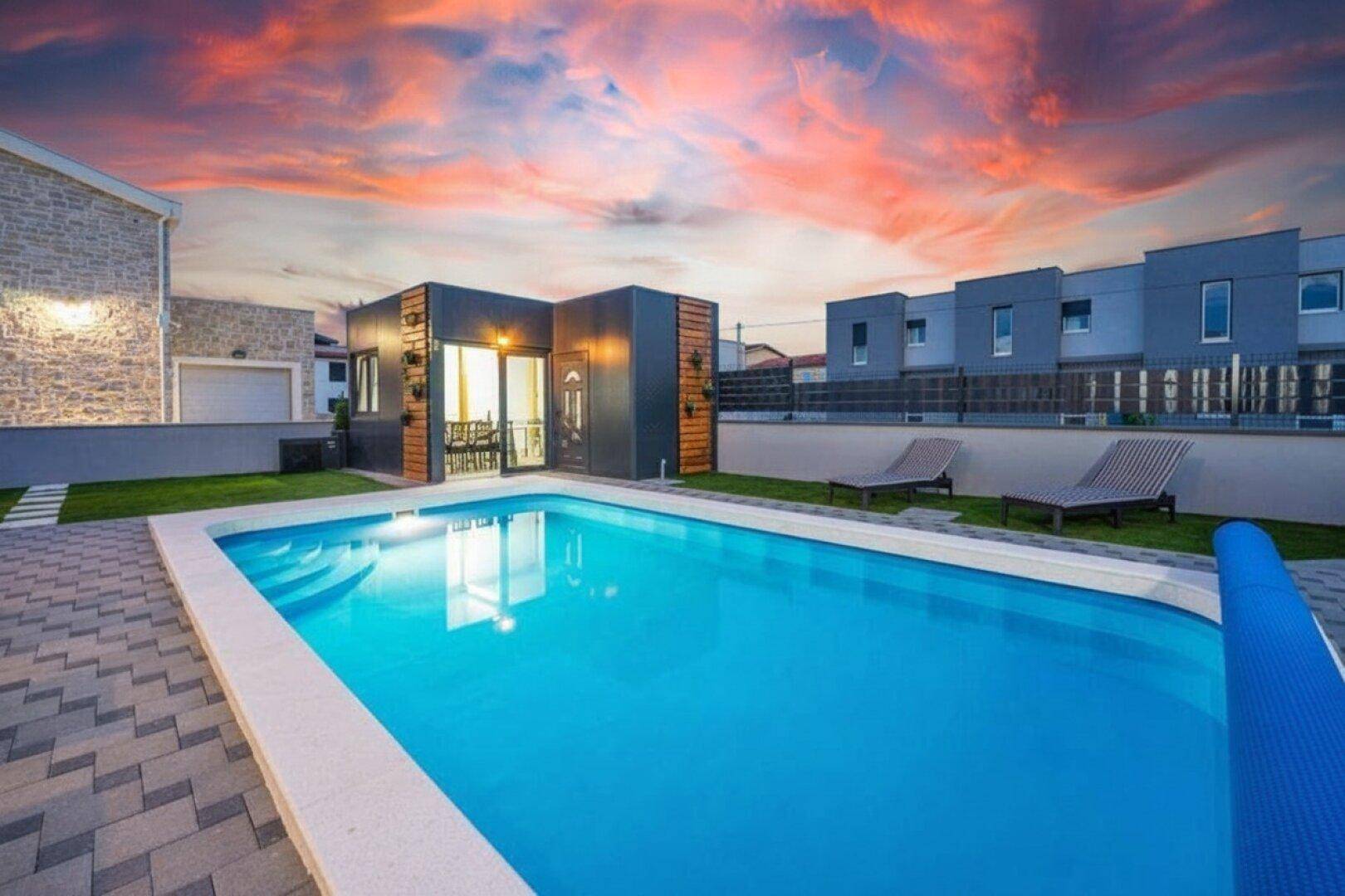 Außenansicht der modernen Villa mit großem Pool und angrenzendem Gebäude unter einem farbenprächtigen Himmel.