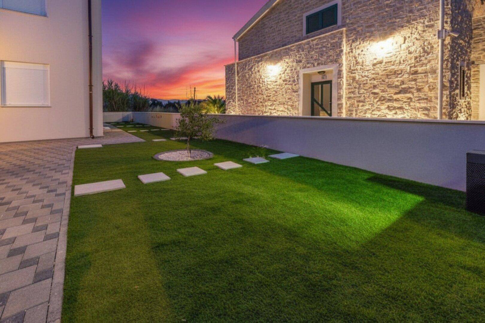 Gepflegter Gartenbereich mit Rasenfläche und Trittsteinen neben dem steinverkleideten Gebäude bei Sonnenuntergang.