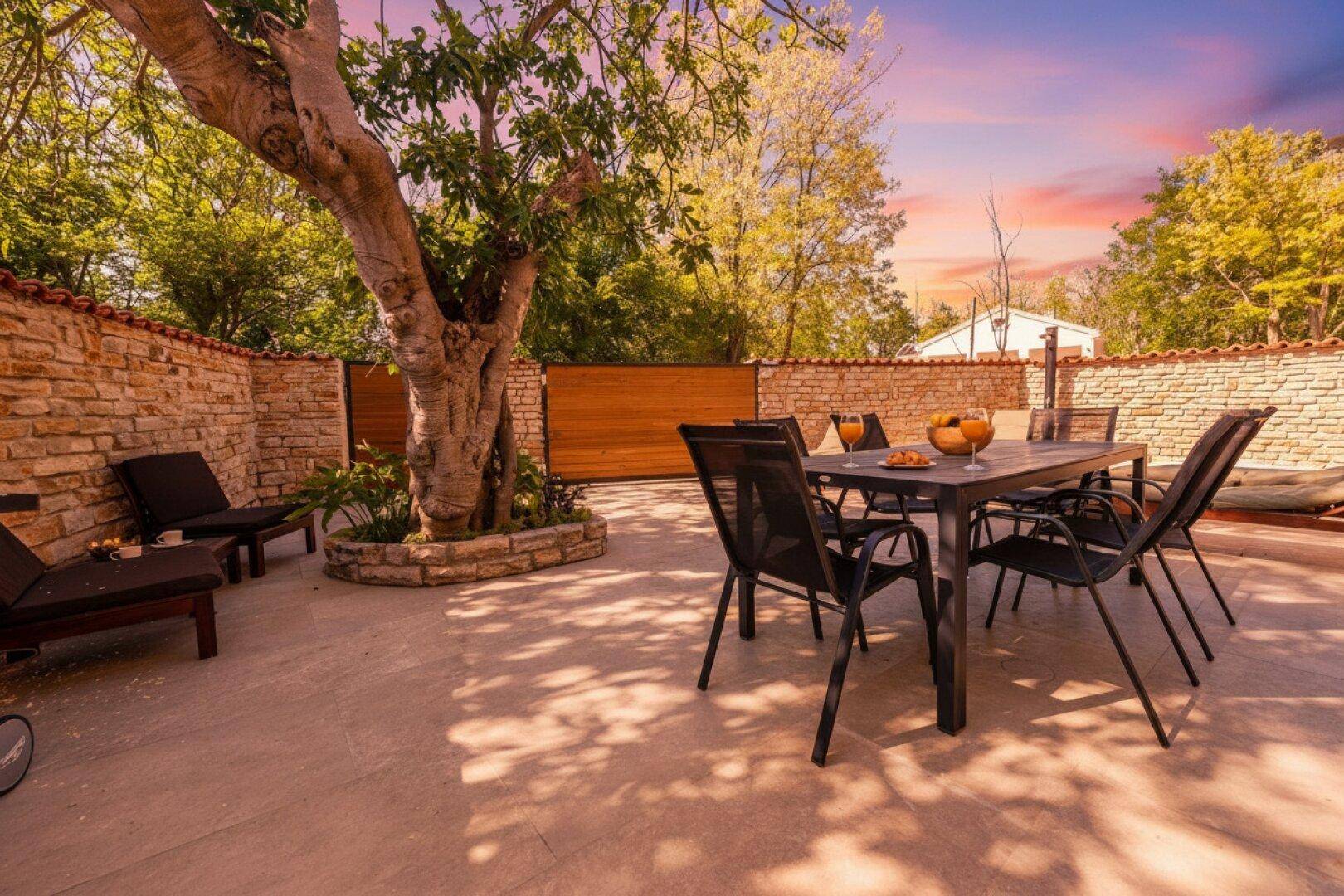 Gemütliche Terrasse mit Esstisch, Liegestühlen und einem schattenspendenden Baum, umgeben von Steinmauern.