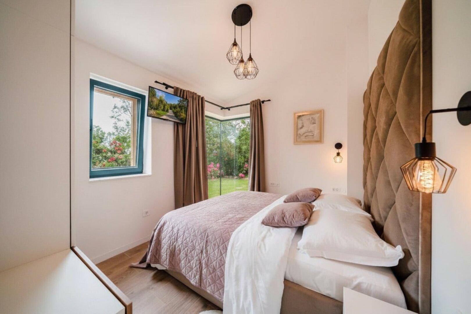 Gemütliches Schlafzimmer mit großem Bett, gepolstertem Kopfteil und Fenstern mit Blick ins Grüne.