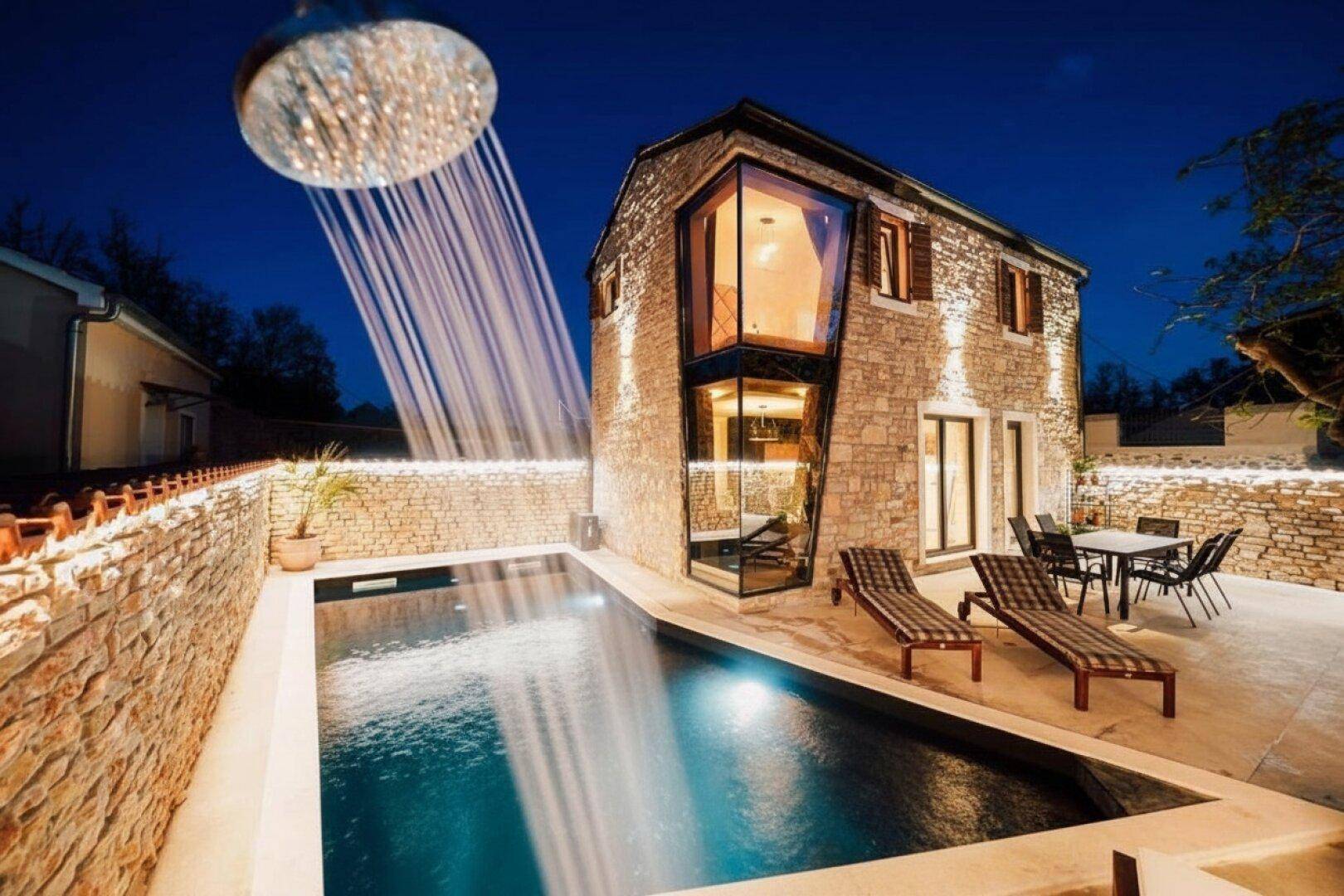 Beleuchtetes Haus mit Pool und Außendusche, umgeben von einer Steinmauer, bei Nacht.