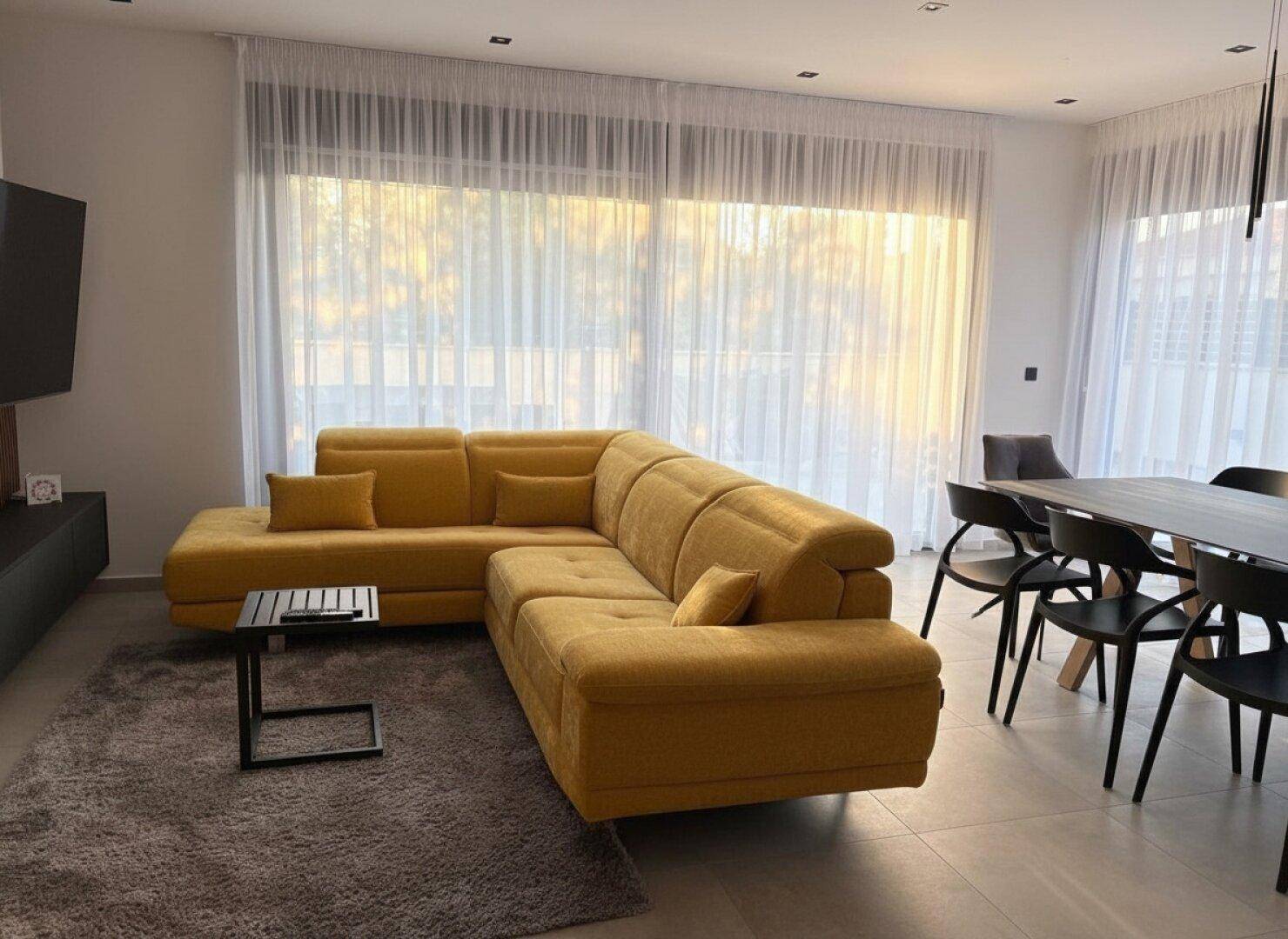Geräumiger Wohnbereich mit bequemem gelbem Ecksofa und Blick auf die großen Fenster.