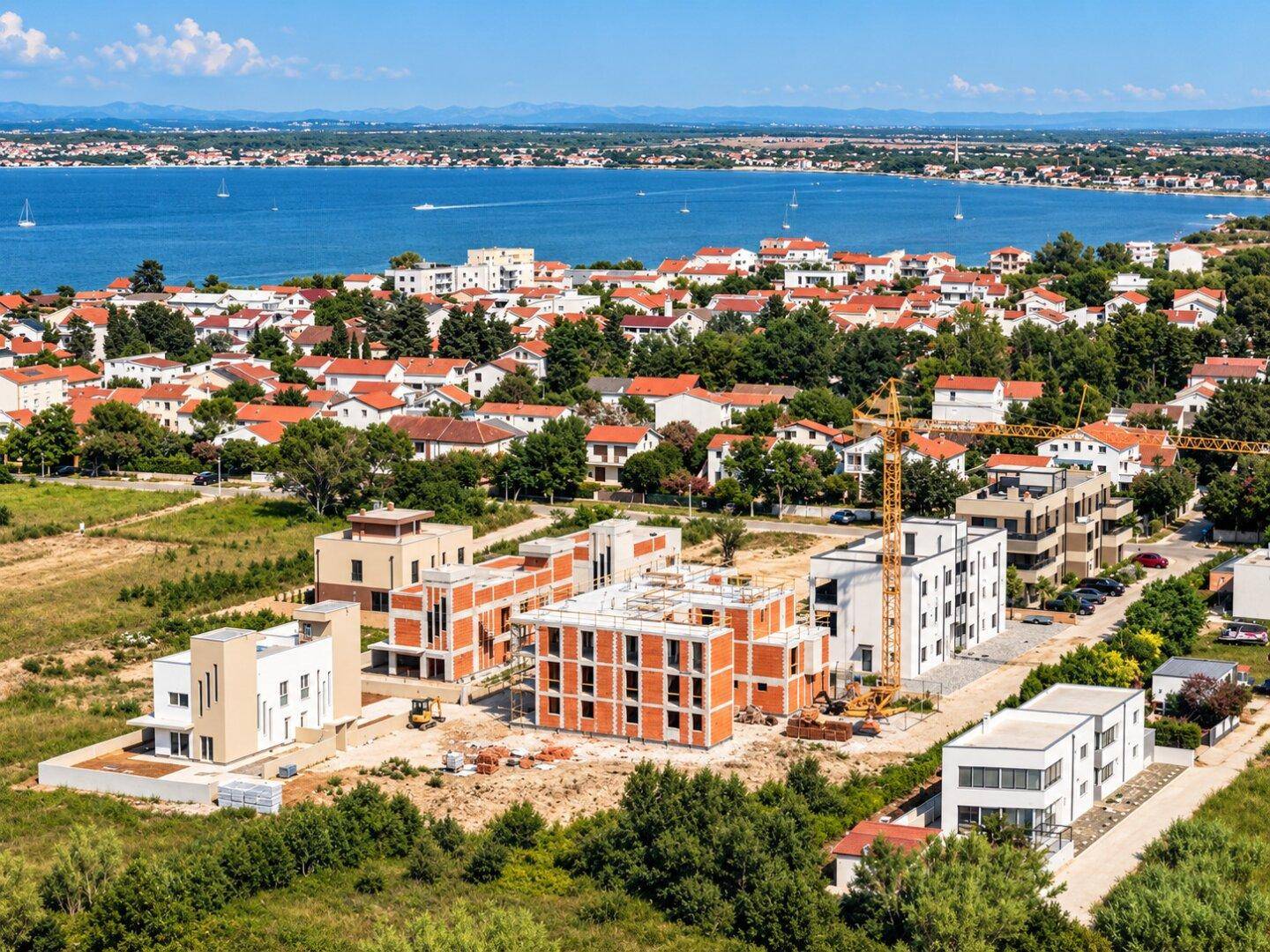 Panoramablick aus der Luft auf eine Küstenstadt mit Neubauten und dem glitzernden Meer im Hintergrund.