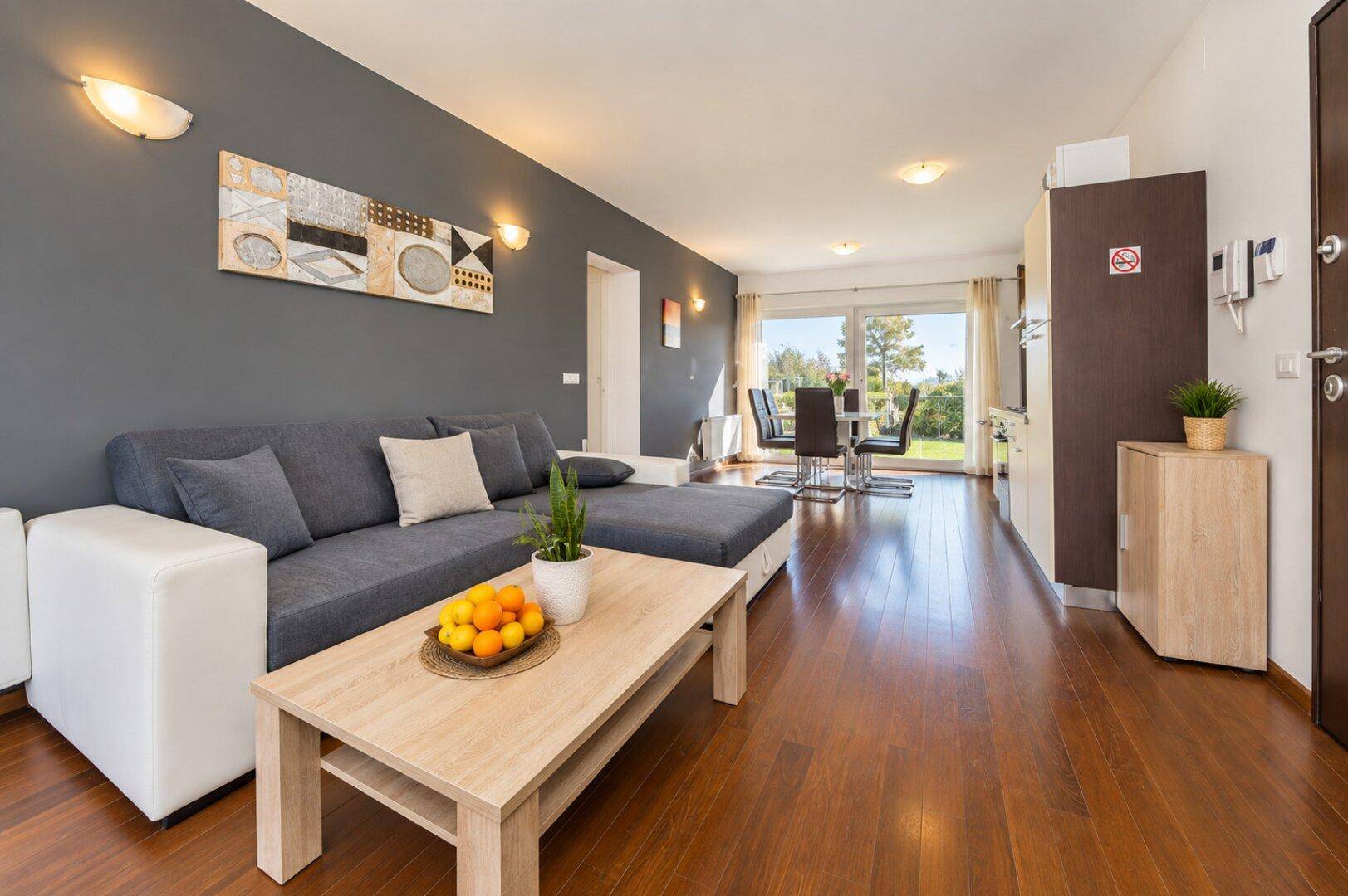 Gemütlicher Wohn- und Essbereich mit dunklem Holzboden, grauem Sofa und Zugang zur Terrasse.